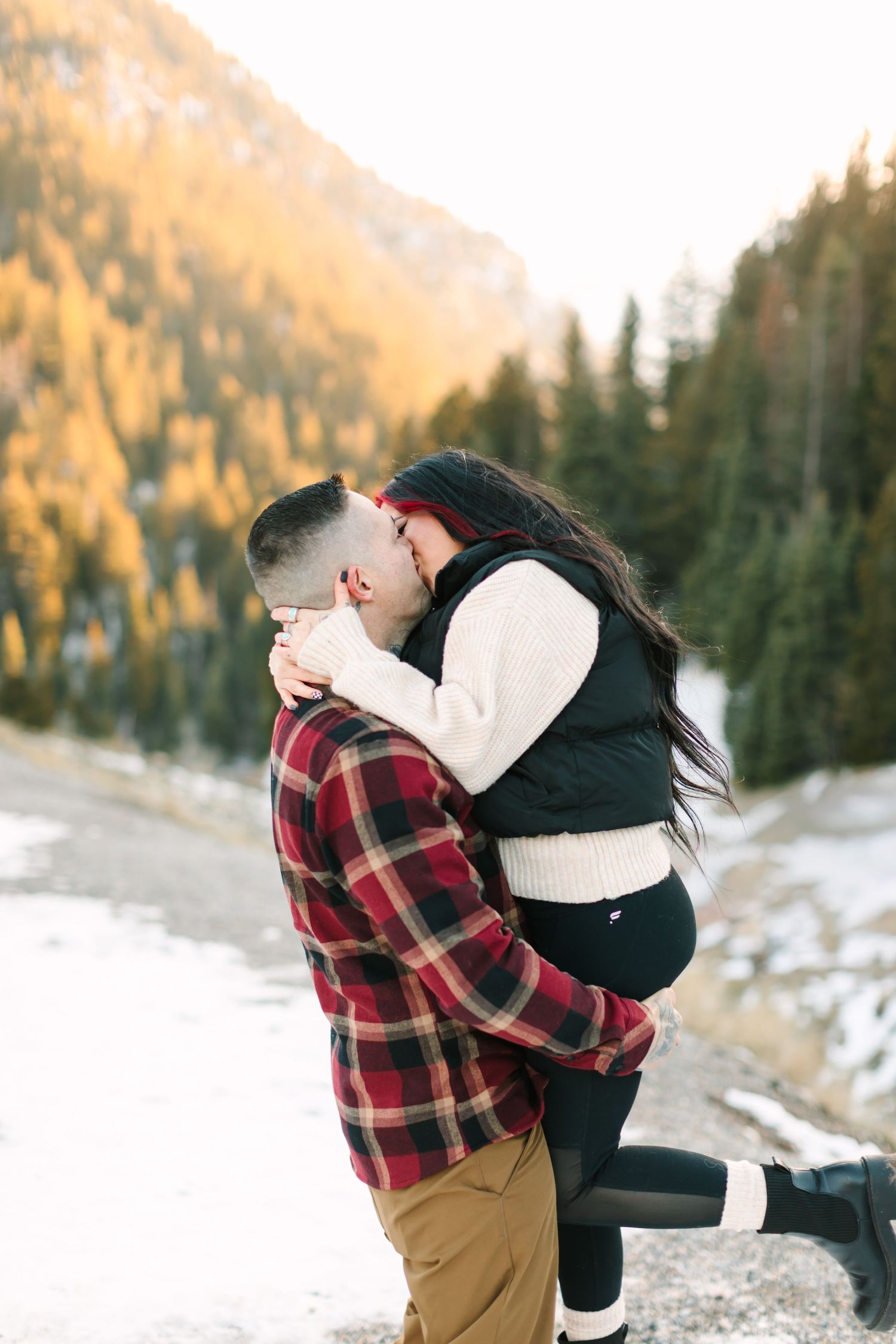A couple shares an intimate moment in a snowy mountain landscape with evergreen trees and golden sunlight.
