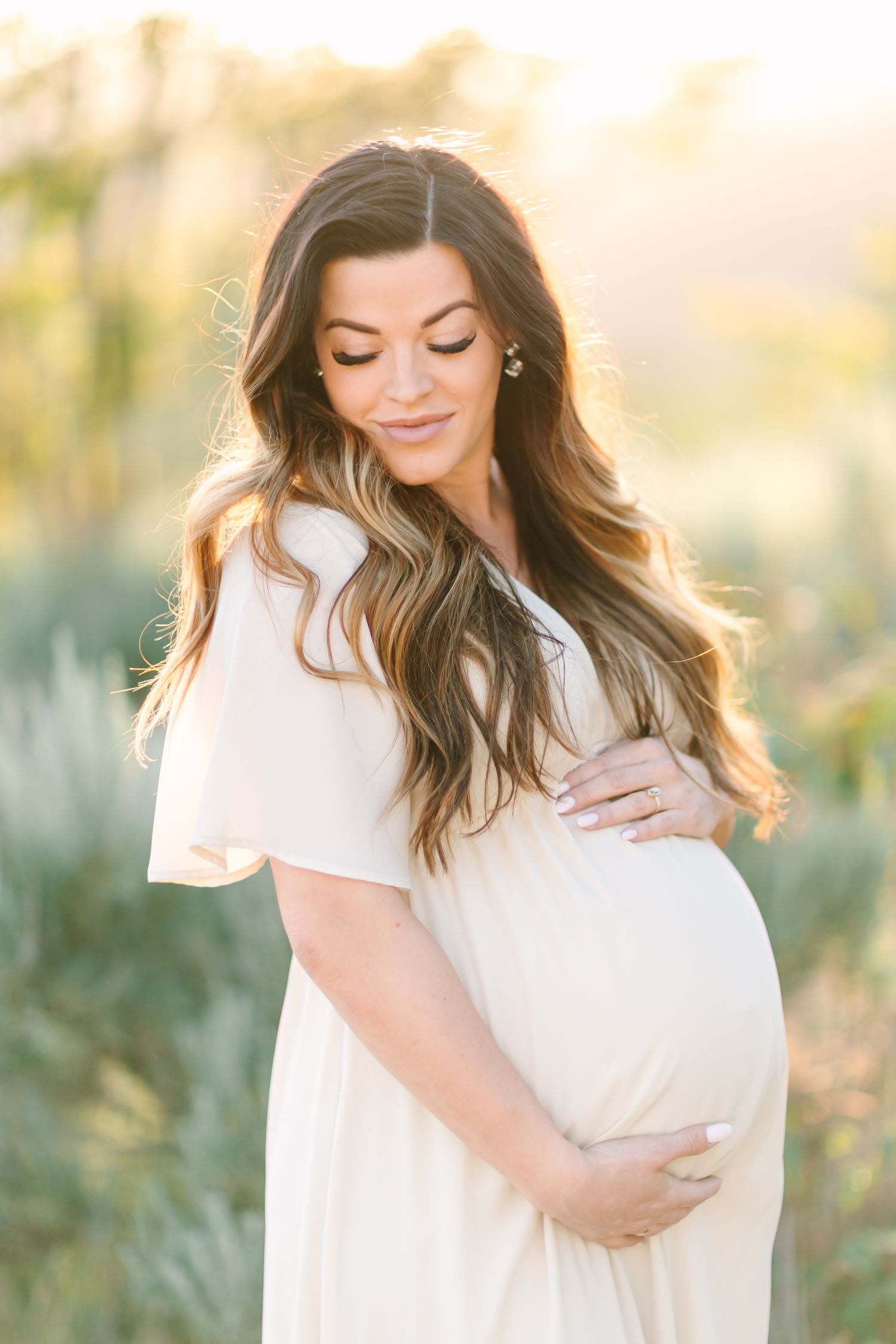 Expectant mother in flowing white dress poses outdoors during golden hour with hands cradling her baby bump.