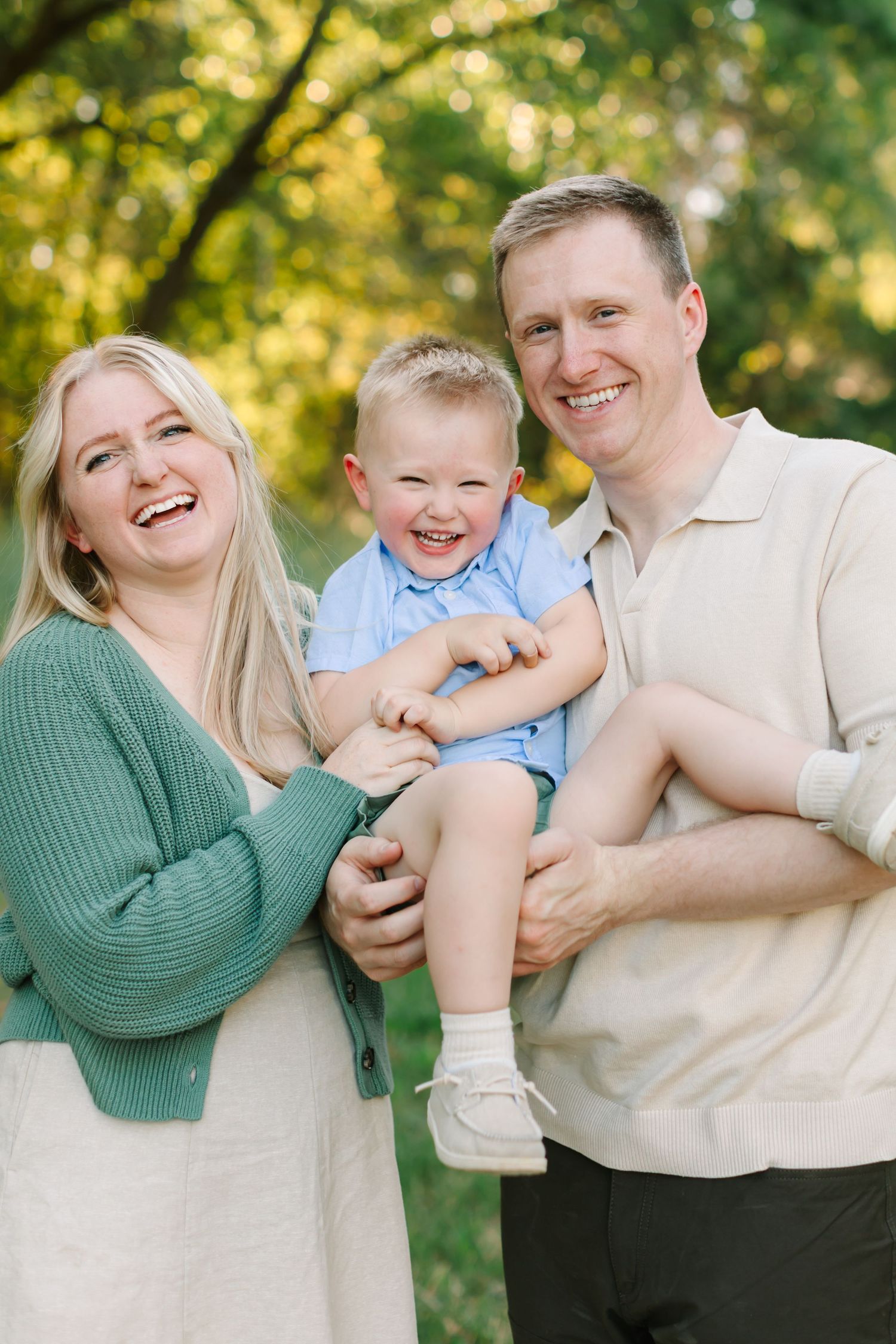 A family shares joyful laughter while playing and embracing outdoors in golden sunlight among green trees.