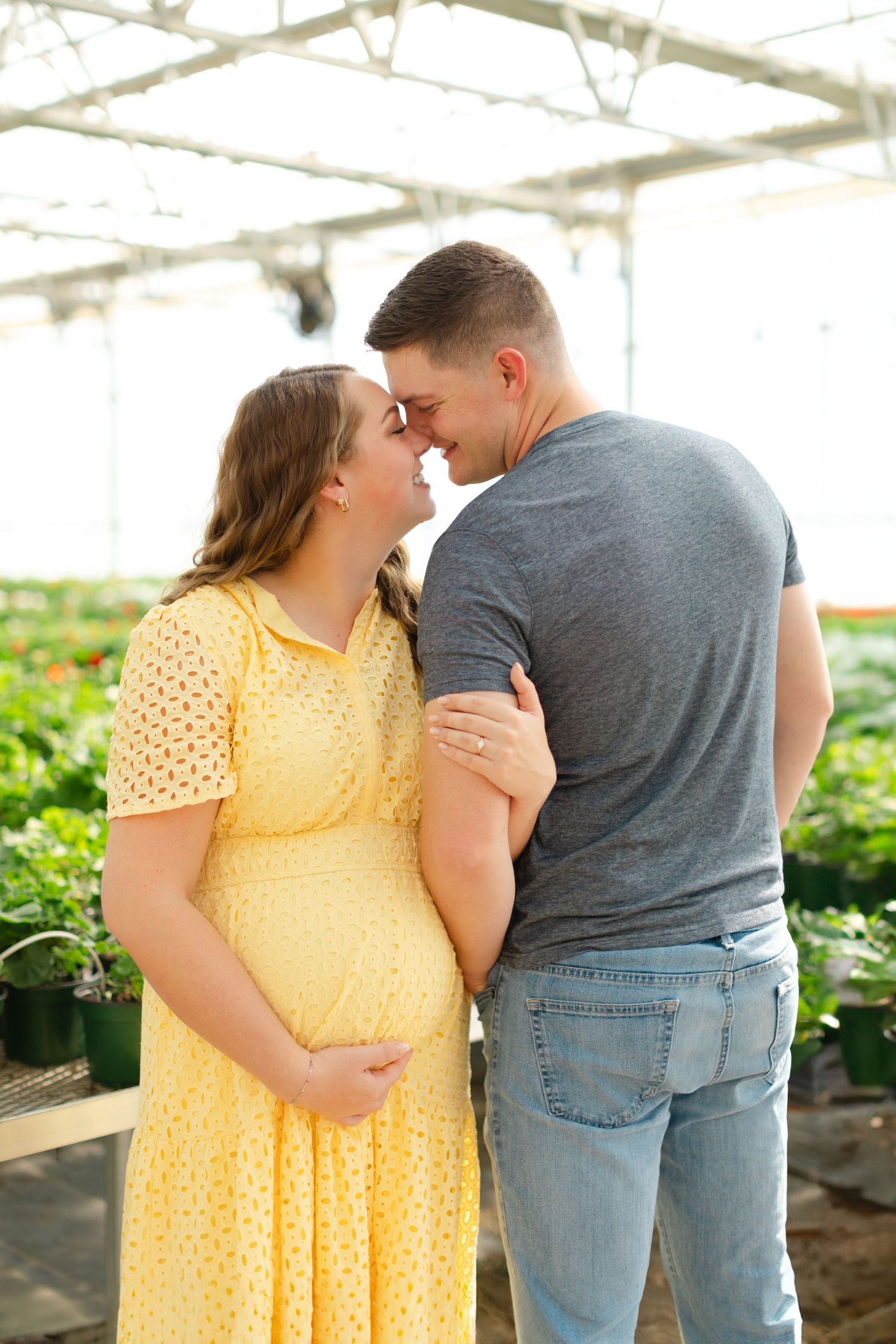 A couple in a greenhouse share an intimate moment surrounded by plants, with one wearing a yellow polka dot dress.
