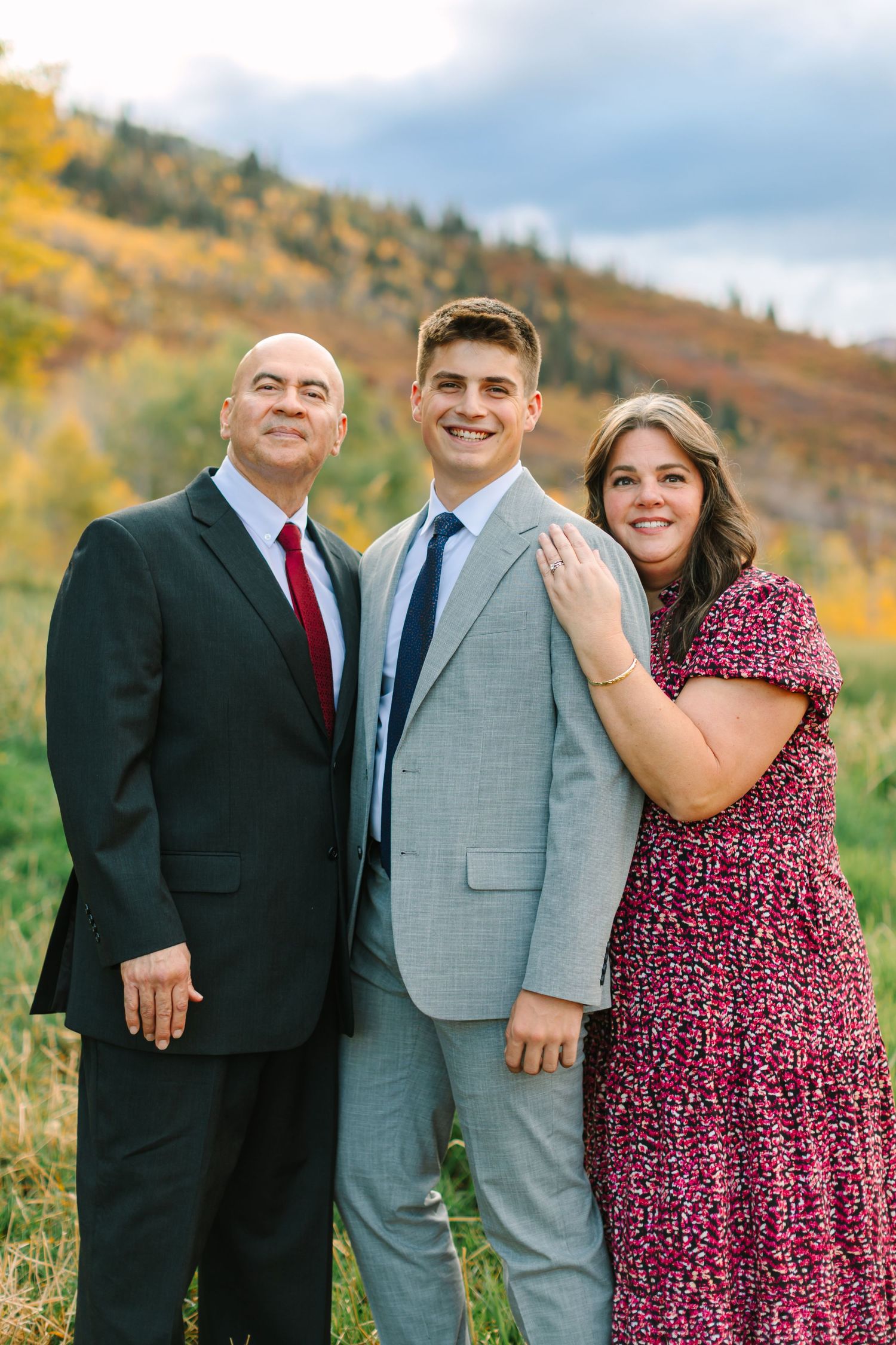 Family portrait of three people in formal attire standing together against a colorful autumn mountain landscape.