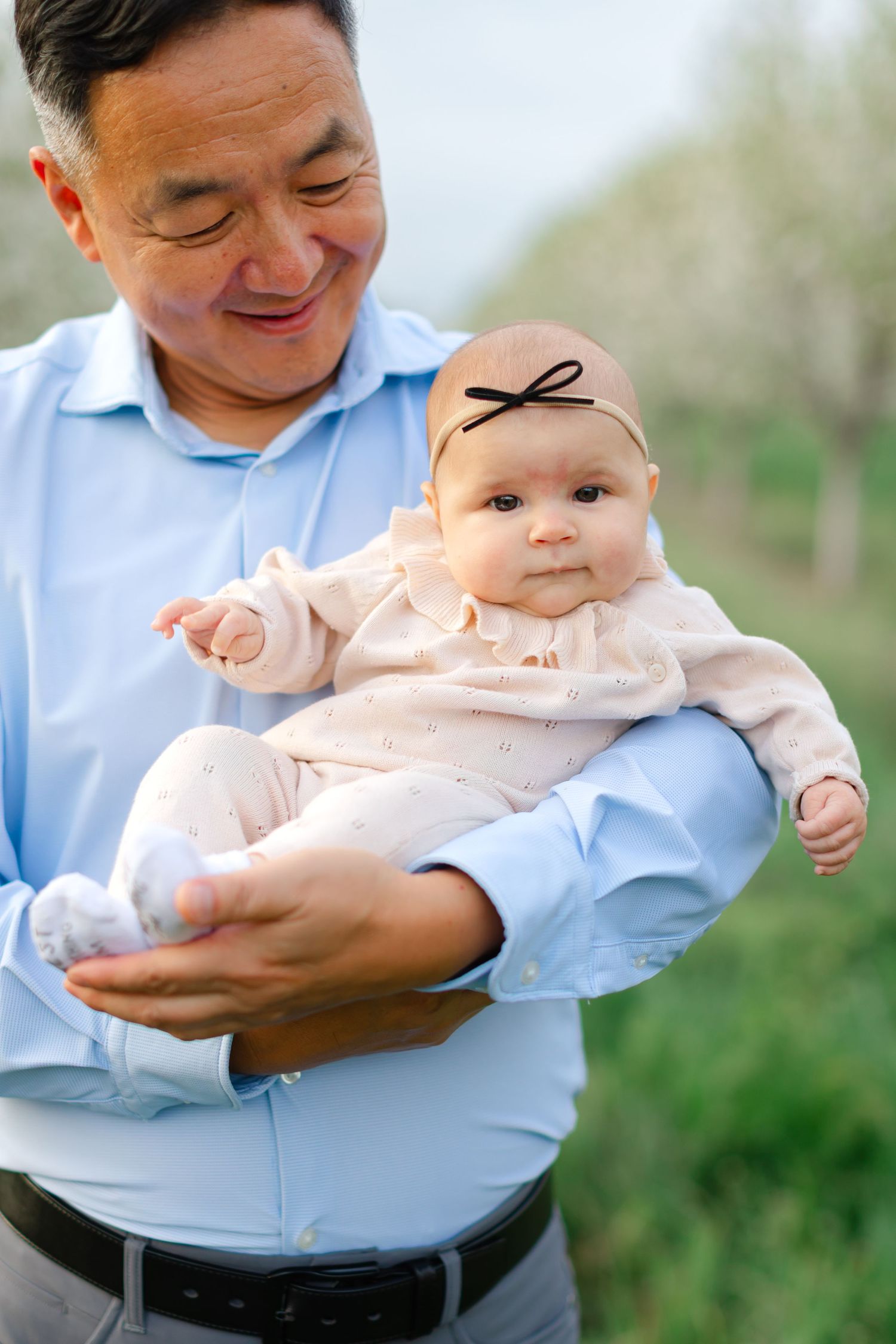 A smiling adult in a light blue shirt holds a baby wearing a pink outfit with a small bow headband outdoors.
