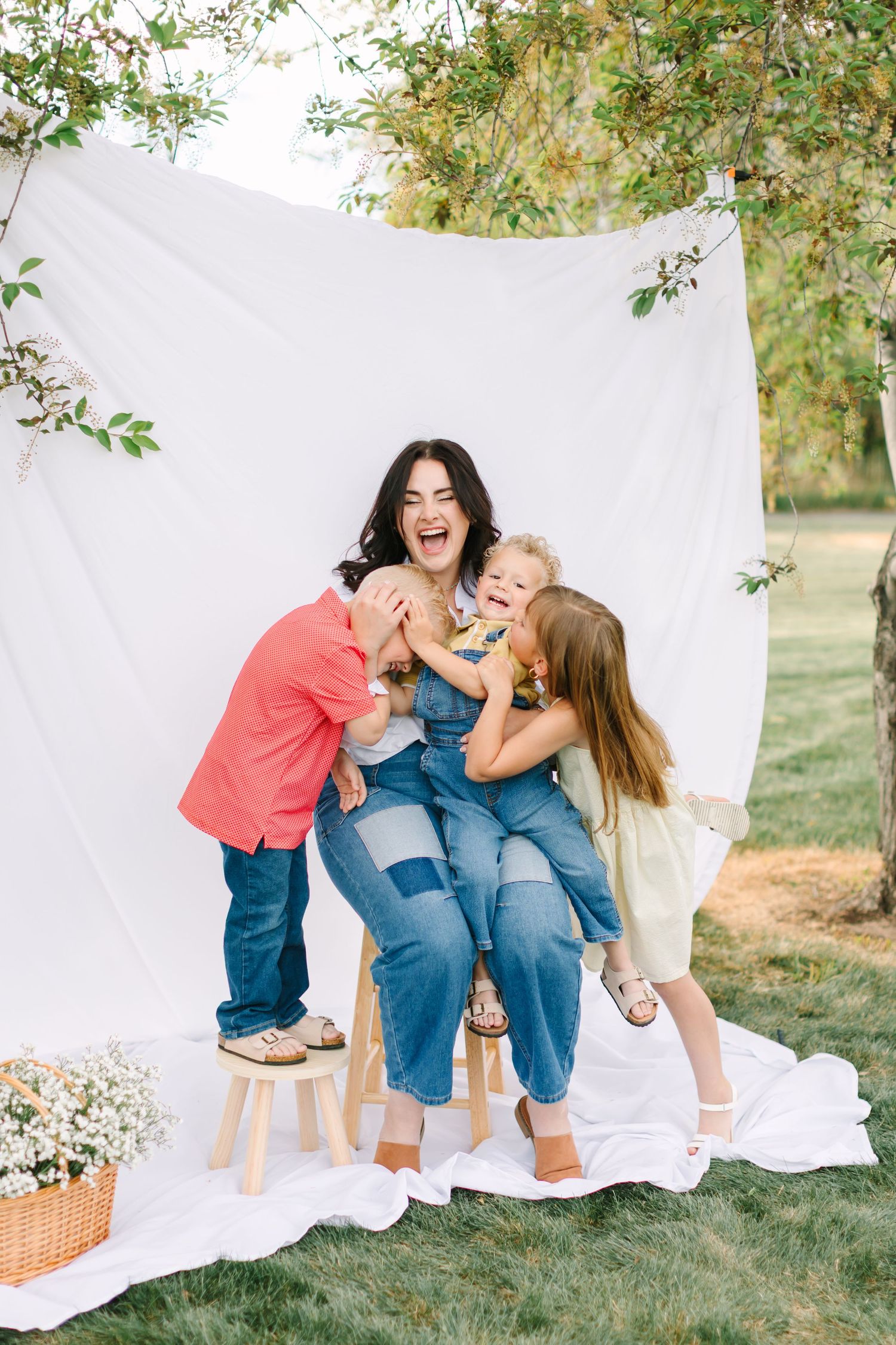 A joyful mother sits outdoors laughing as her three children playfully hug and kiss her during a family photo session.