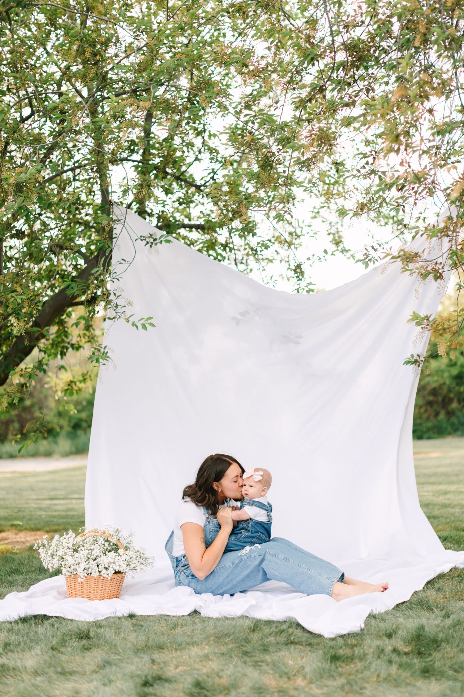 A mother sits on a white blanket outdoors, kissing her baby with a white fabric backdrop and flower basket nearby.
