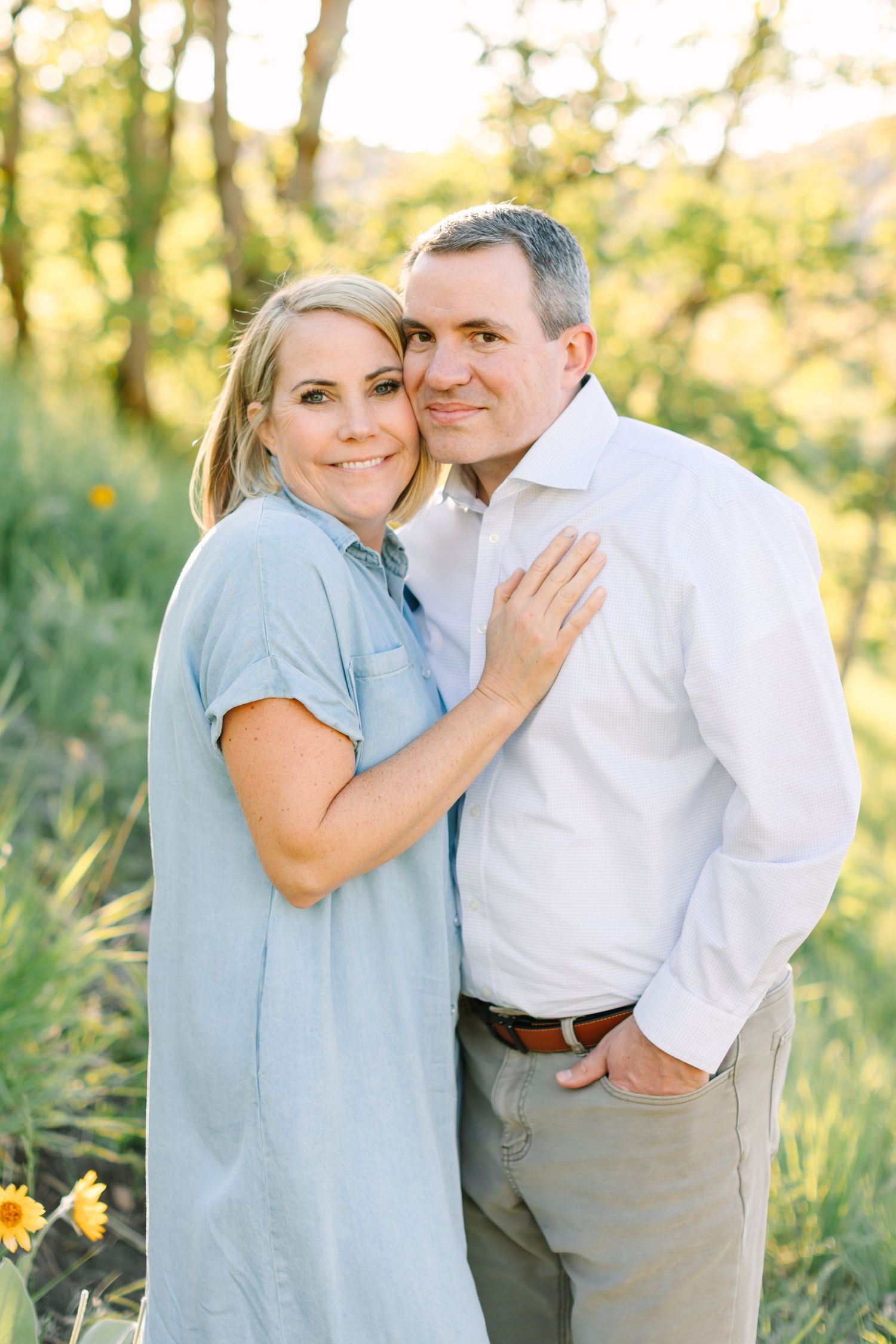 A smiling couple poses outdoors in golden light, woman in blue dress leaning on man in white shirt amid green foliage.