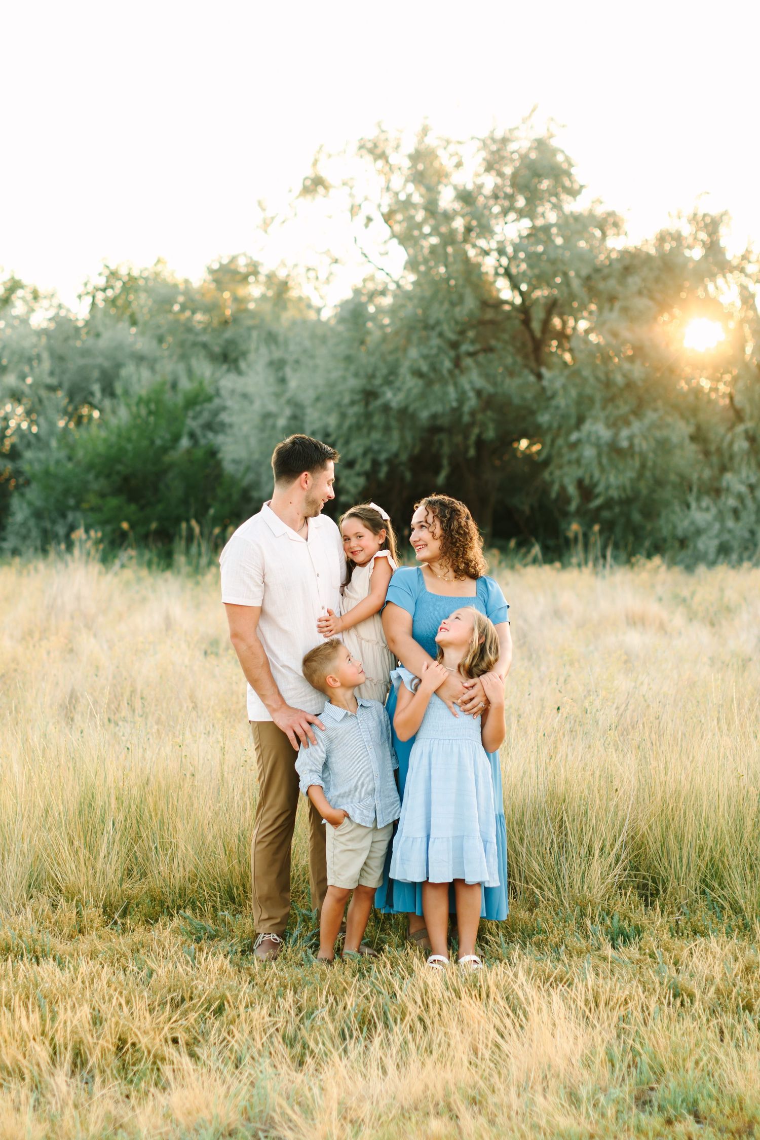 Family of five posing in golden field at sunset, parents holding children in blue outfits with trees in background in Bountiful, Utah.