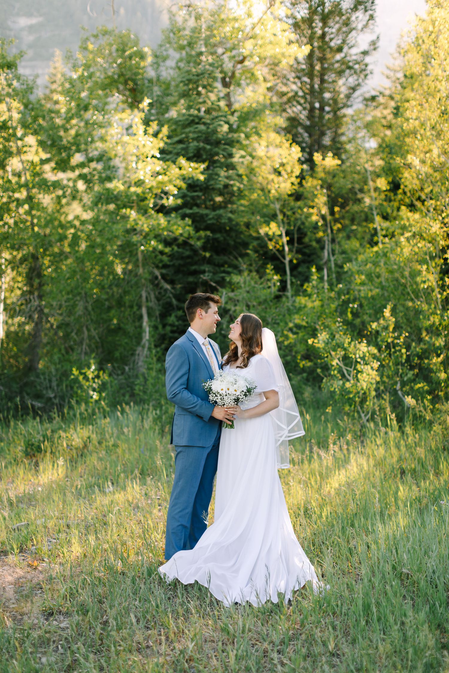 Bride and groom share a romantic moment in a lush green forest meadow, bride holding white bouquet in flowing gown.
