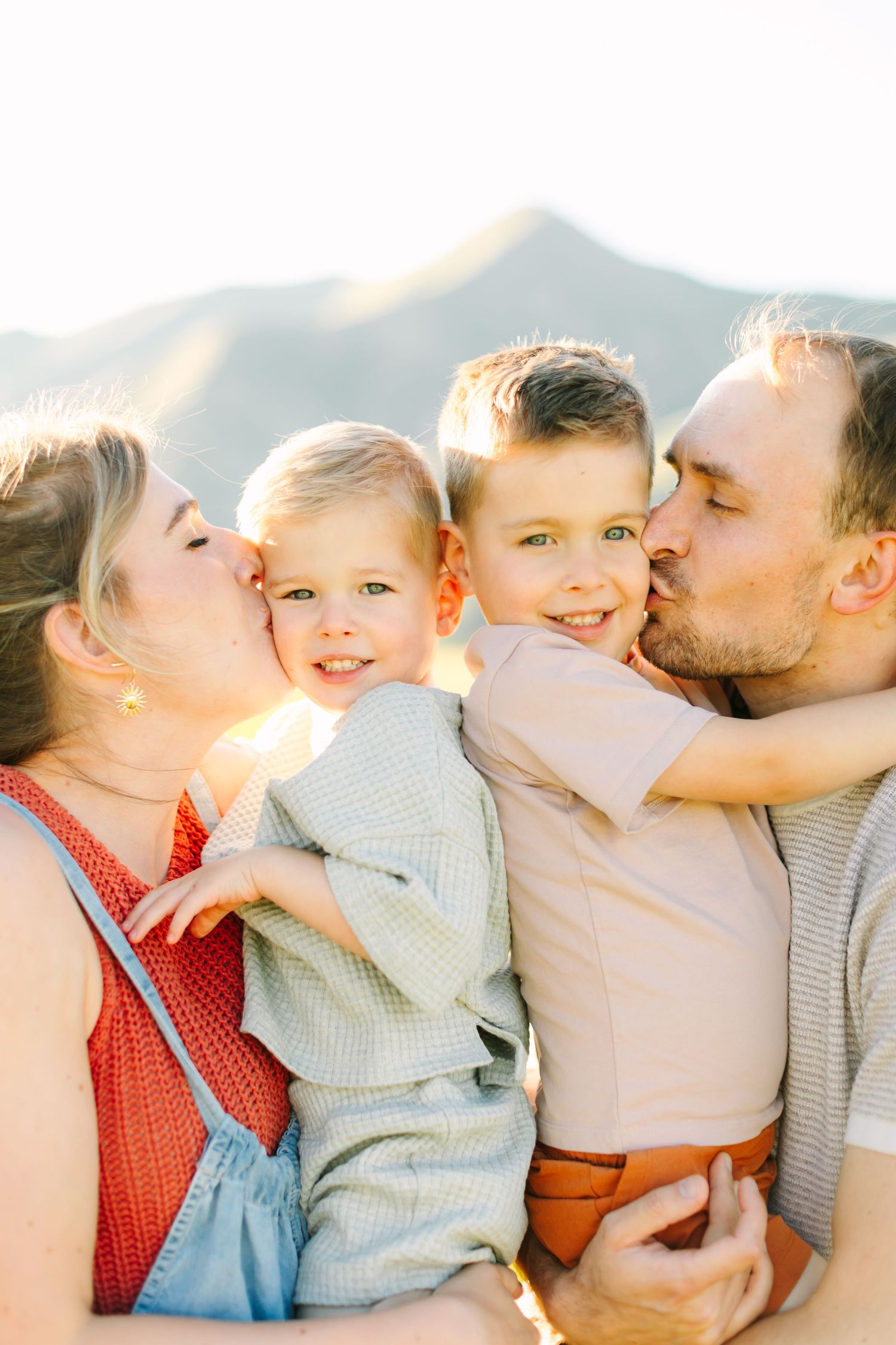 Parents kissing their two young sons on the cheeks outdoors with mountains in the background on a sunny day.