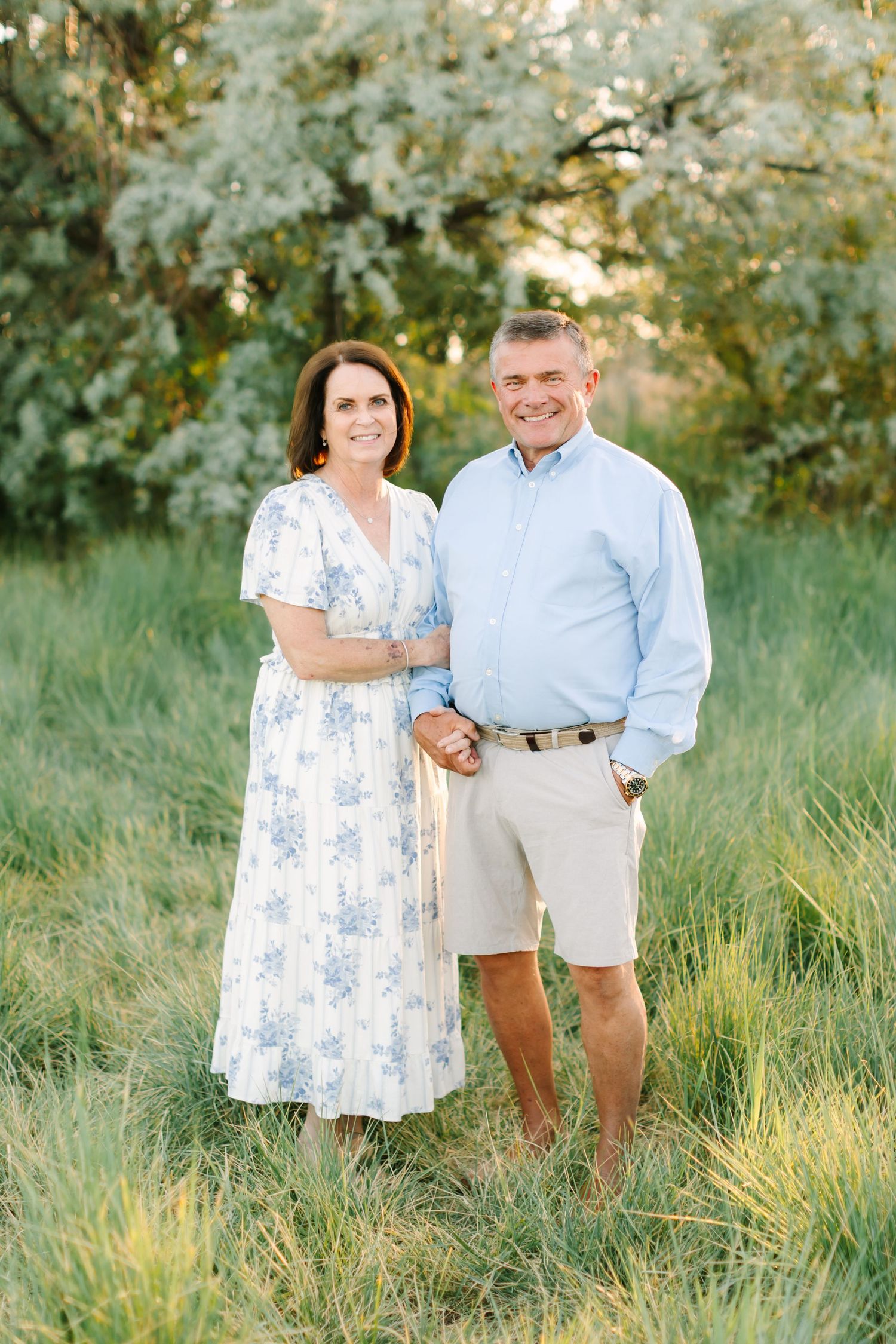 Smiling couple standing in a lush green field, woman in floral dress and man in light blue shirt and khaki shorts.