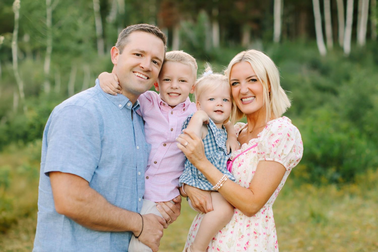 Happy family of four smiling outdoors among green trees, parents holding two young children in a sunny forest setting.