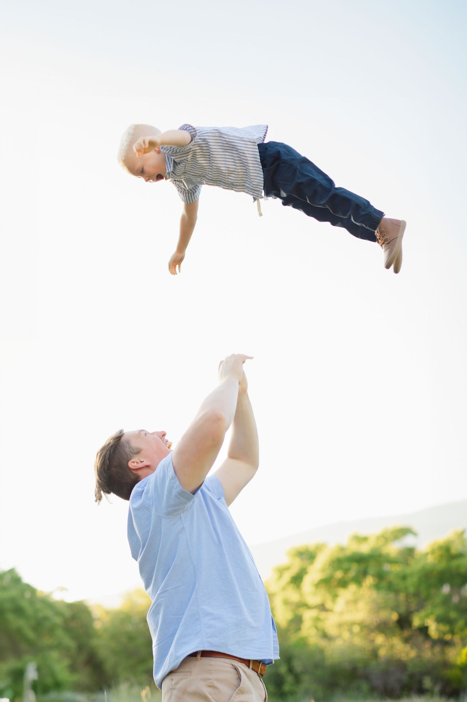 Parent holds toddler up in the air against bright sky and trees during a joyful outdoor playtime moment.