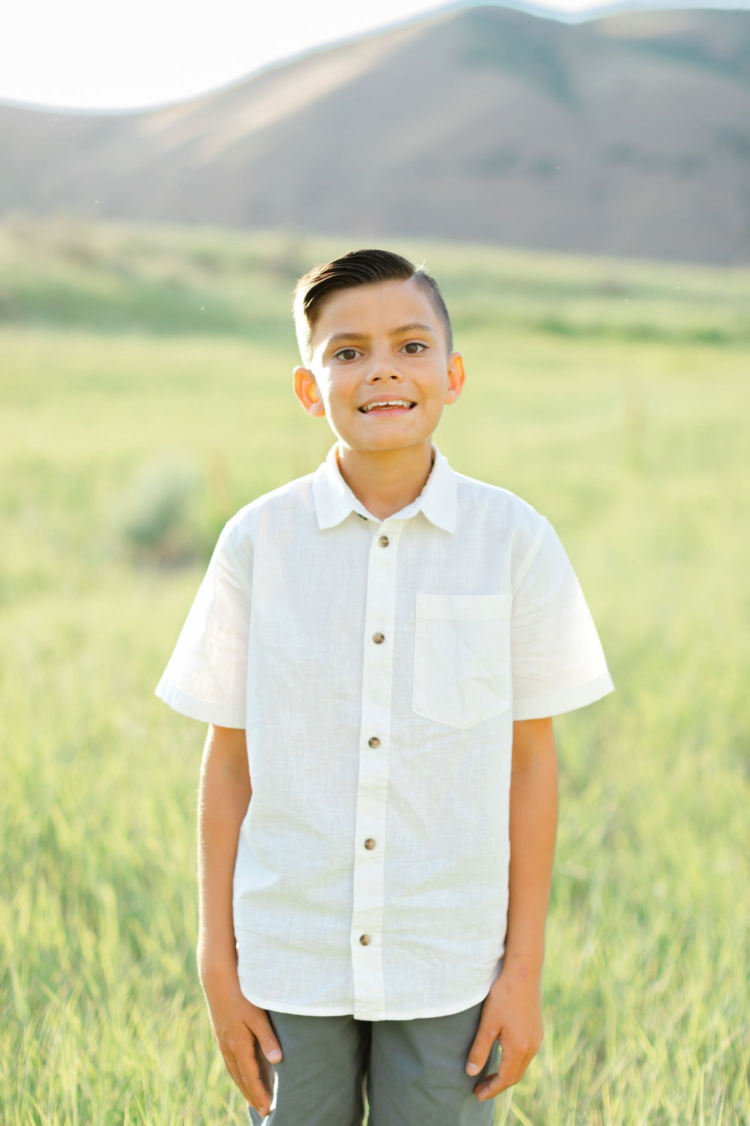 A young person in a white button-down shirt stands smiling in a grassy field with rolling hills in the background.