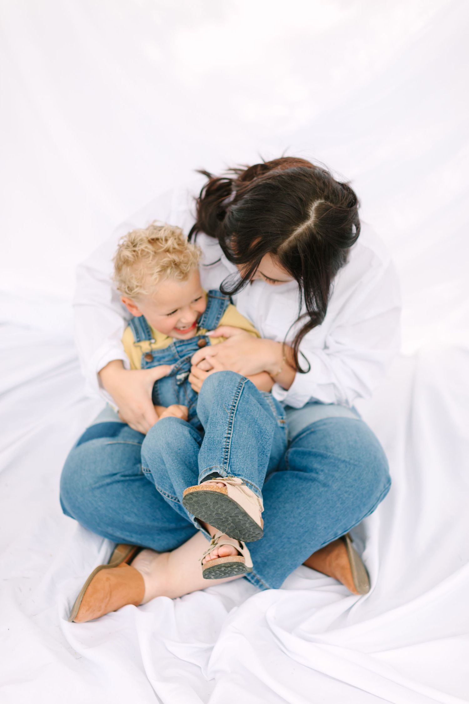 A mother in jeans sits cross-legged on white fabric, tenderly holding her curly-haired toddler in denim overalls.