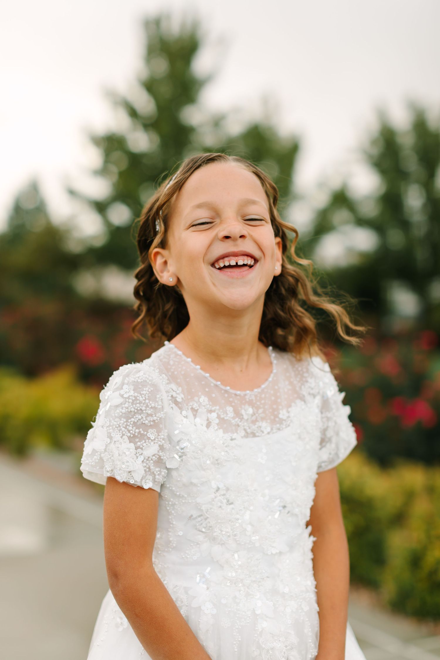 A joyful child wearing a white lace dress laughs and plays outdoors in a garden setting with trees and roses.