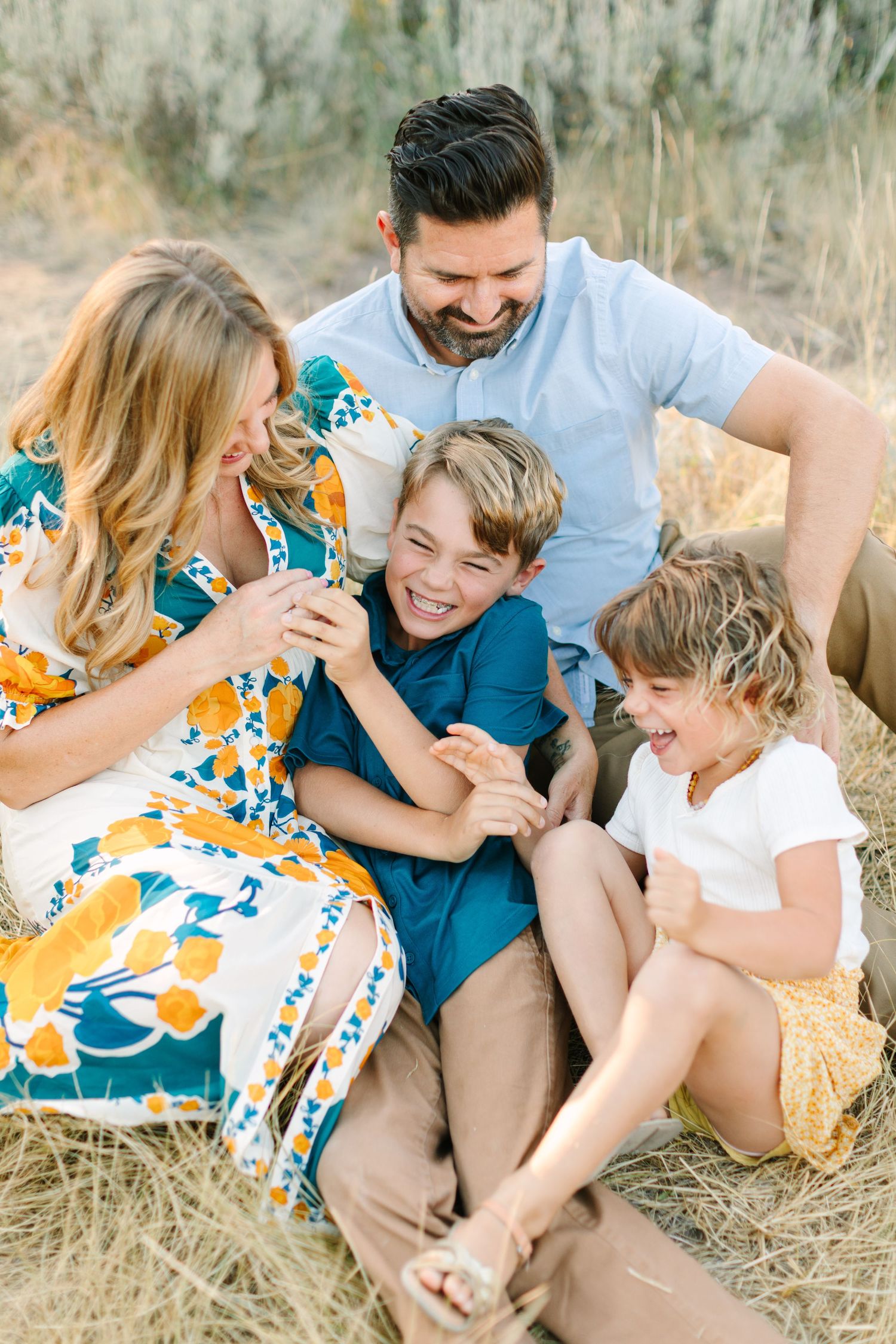 A family laughs together while sitting in a grassy field during a casual outdoor photo session.