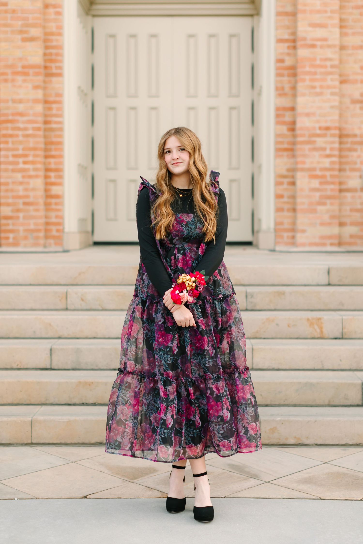 Woman in black and pink floral dress poses elegantly on stone steps against brick wall backdrop.