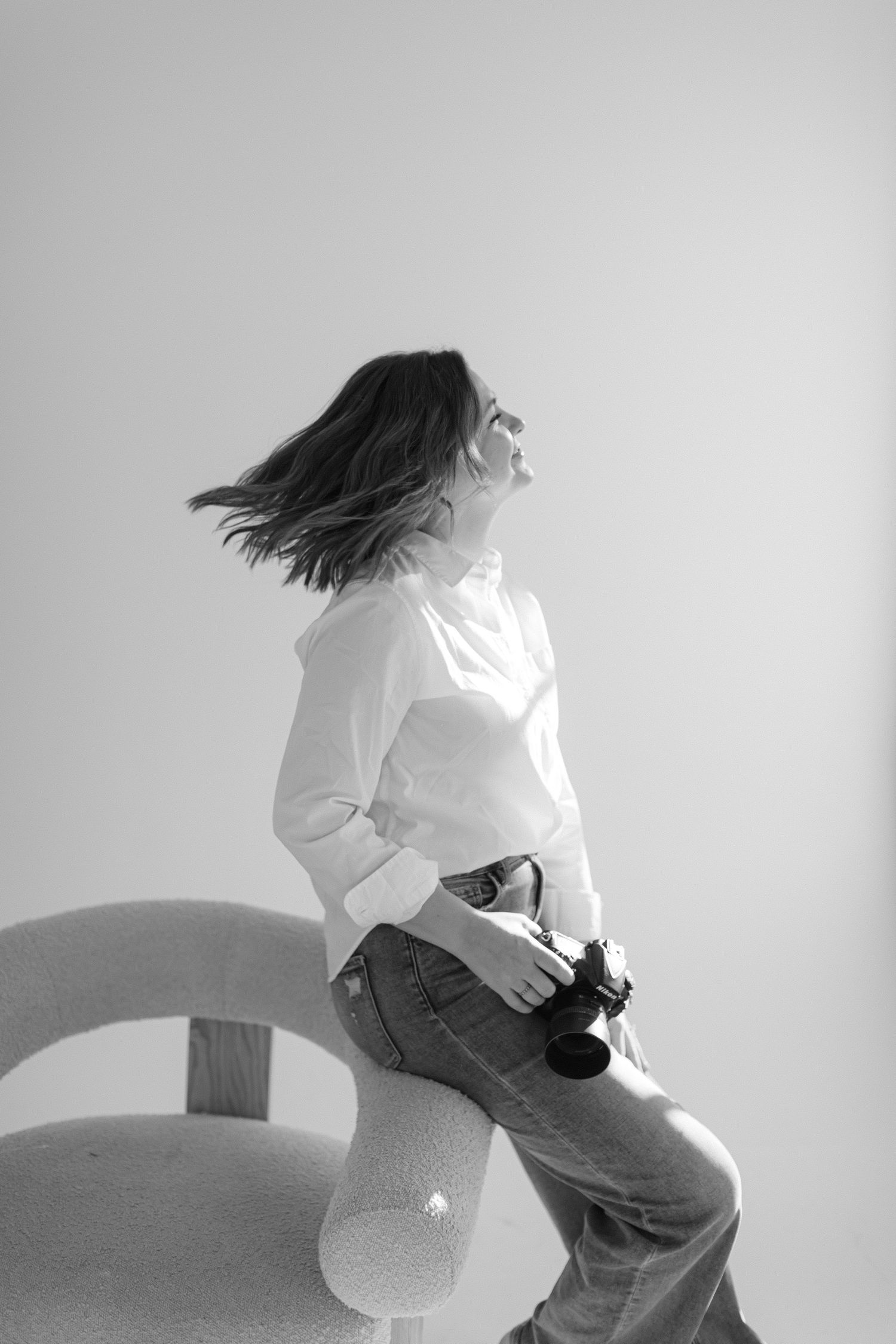 Black and white photo of someone in a white top and jeans sitting on furniture with hair in motion against light background.