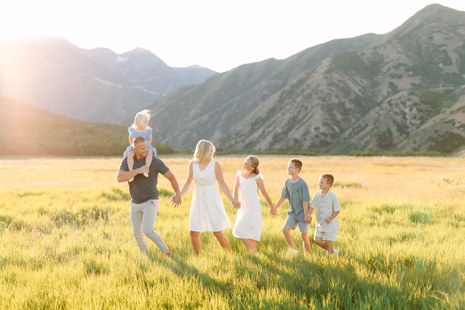 Family walking in sunlit mountain meadow, parents with four children holding hands at golden hour.