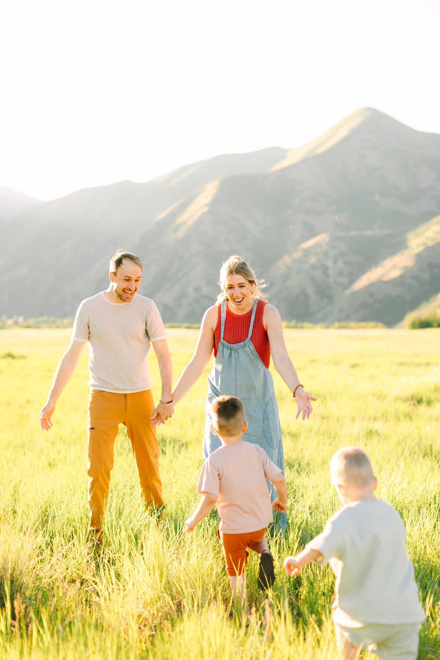 Happy family with two boys playing in a sunny green field with mountains in the background.