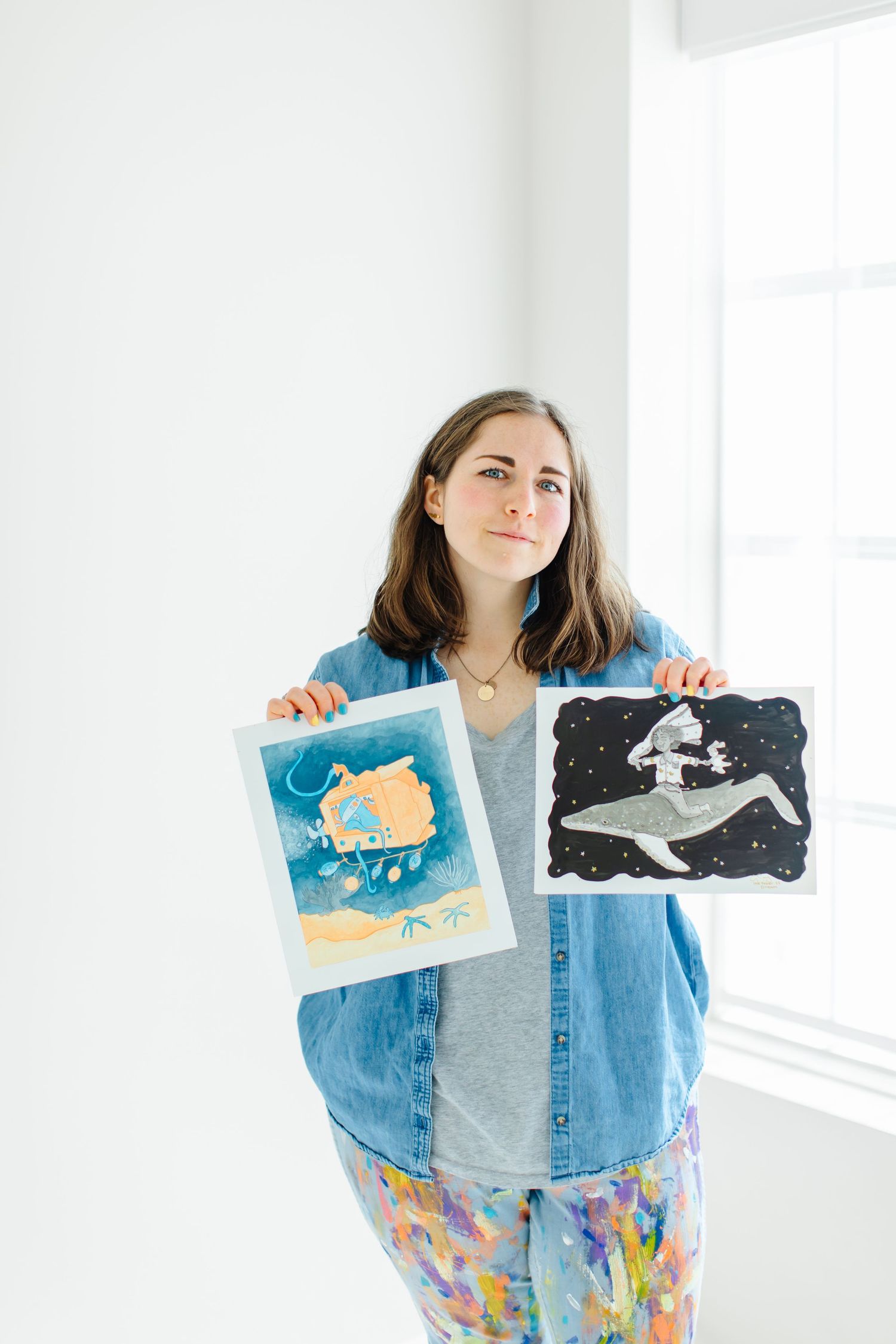 Artist displays two artwork pieces while wearing colorful pants and denim shirt at White Space Studios in Provo, Utah in Utah County..