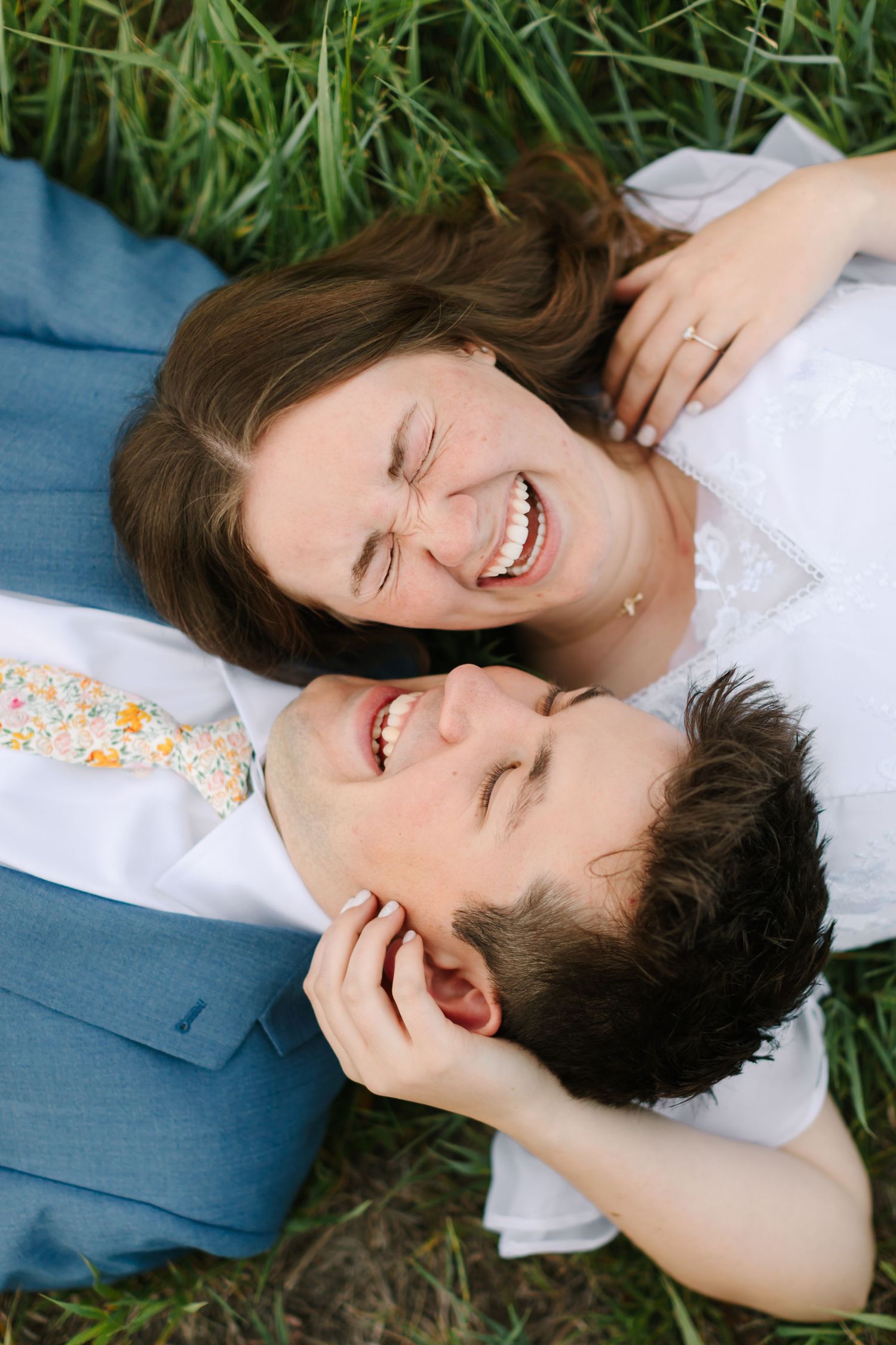 Happy couple laughing together while lying in green grass, bride in white dress and groom in blue suit with floral tie.