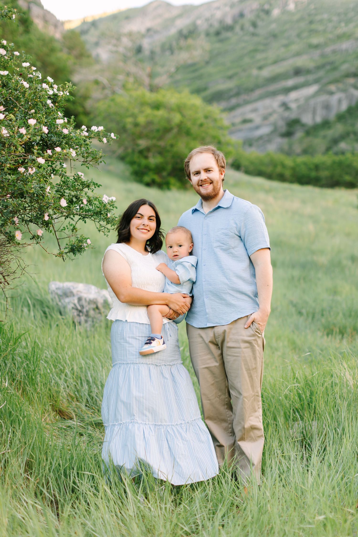 A young family with a baby poses in a lush green mountain meadow near blooming shrubs during golden hour.
