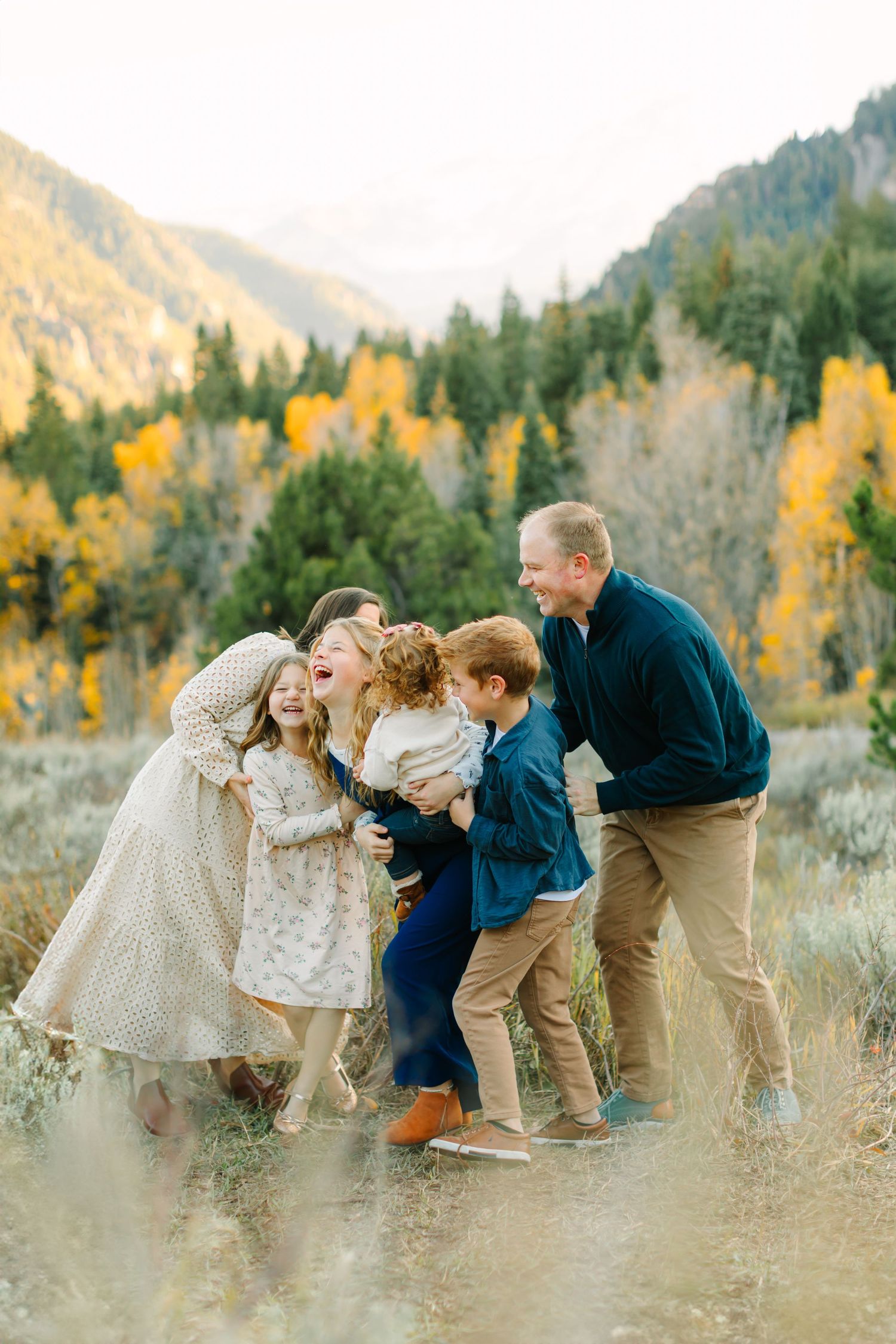 A family in coordinated neutral outfits plays together in a field with autumn mountains and golden aspens in the background.