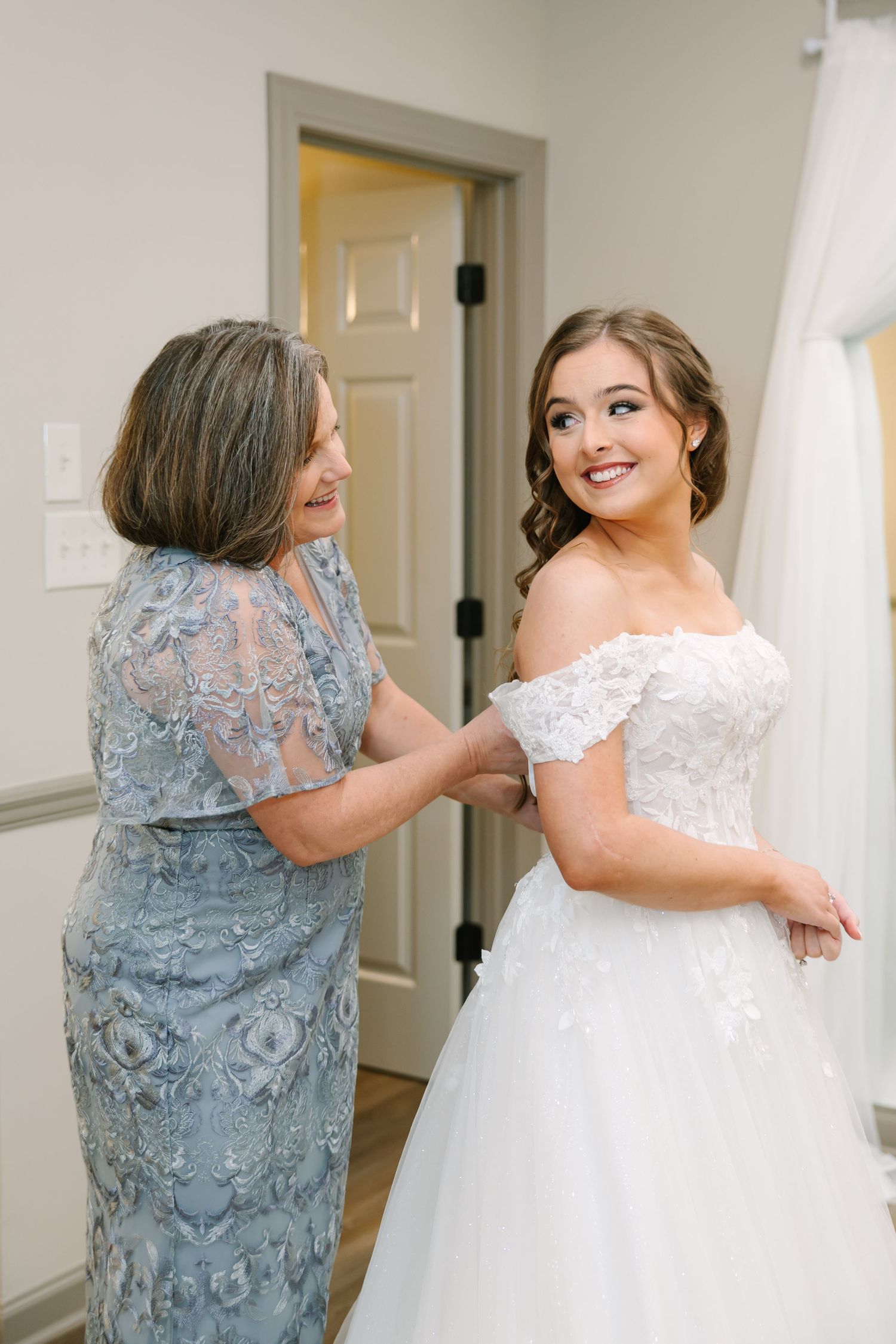 A tender moment between two women as they prepare in formal attire, one in a blue lace gown and one in white wedding dress.
