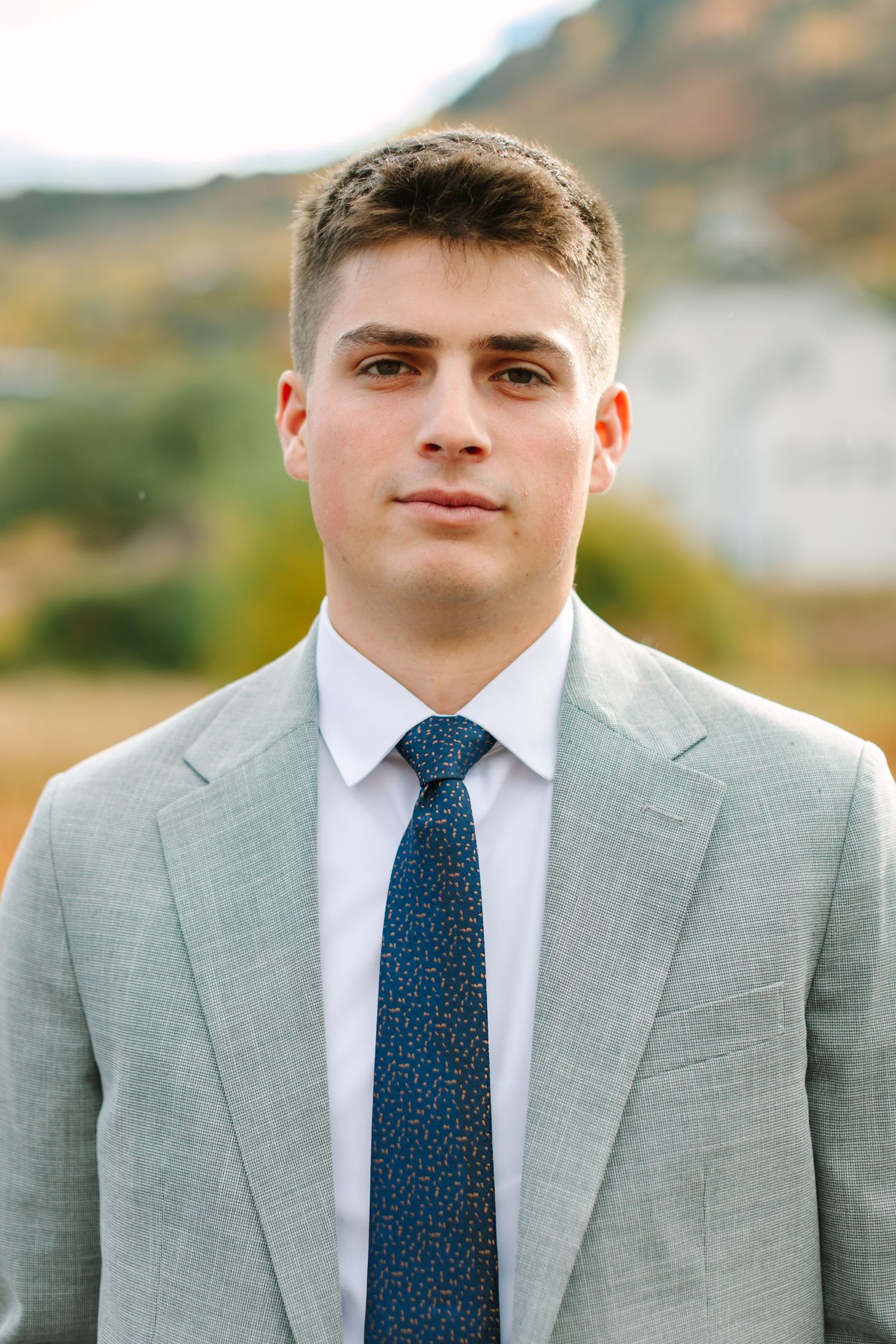 Professional portrait of a young person in a light gray suit with a patterned blue tie against a blurred outdoor background.