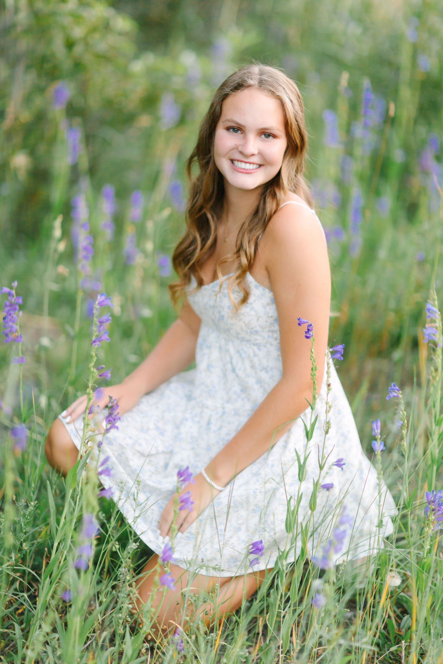 A person in a white lace dress poses in a field of purple wildflowers on a sunny summer day.