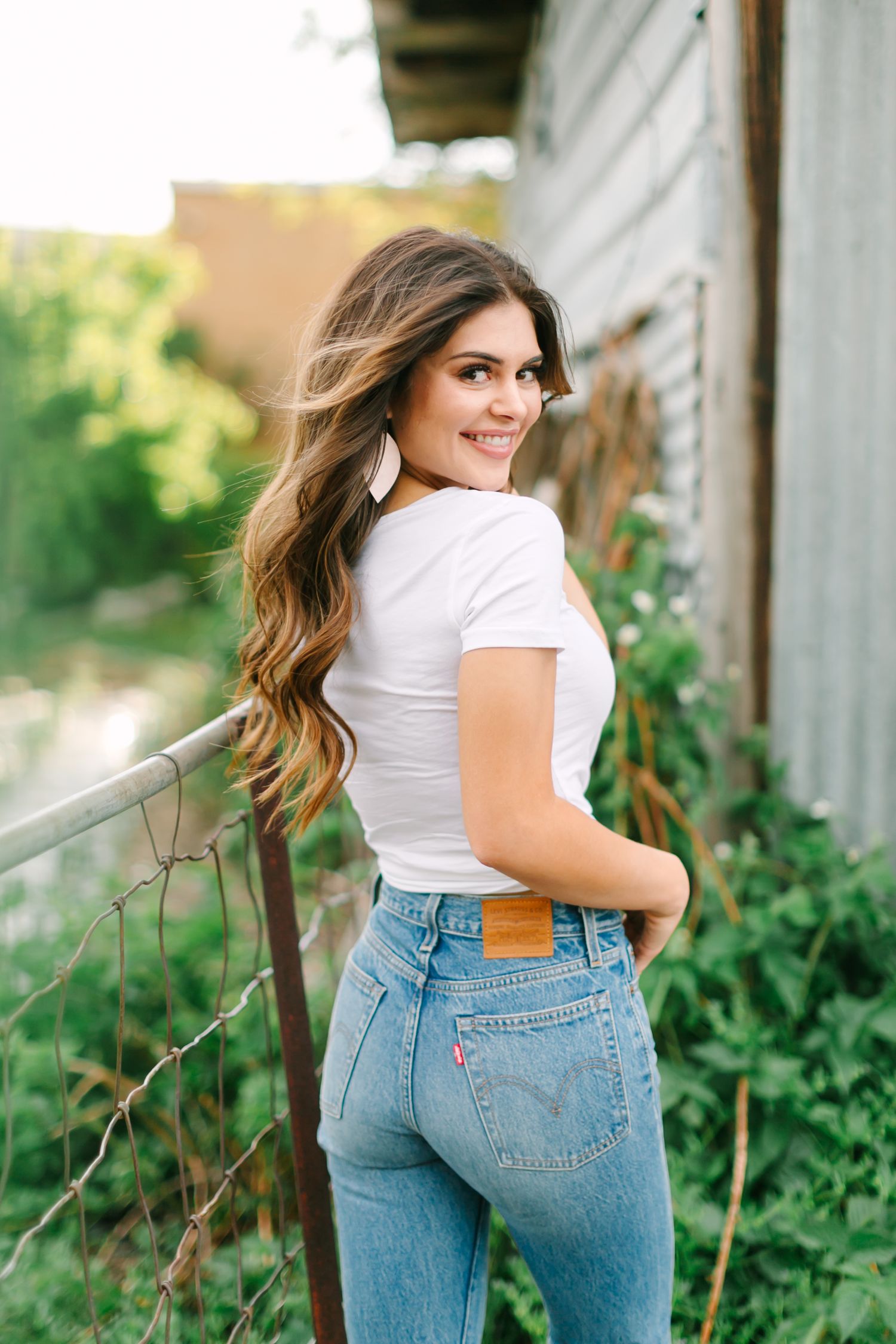 Woman in white t-shirt and high-waisted jeans poses outdoors against rustic wall with greenery.