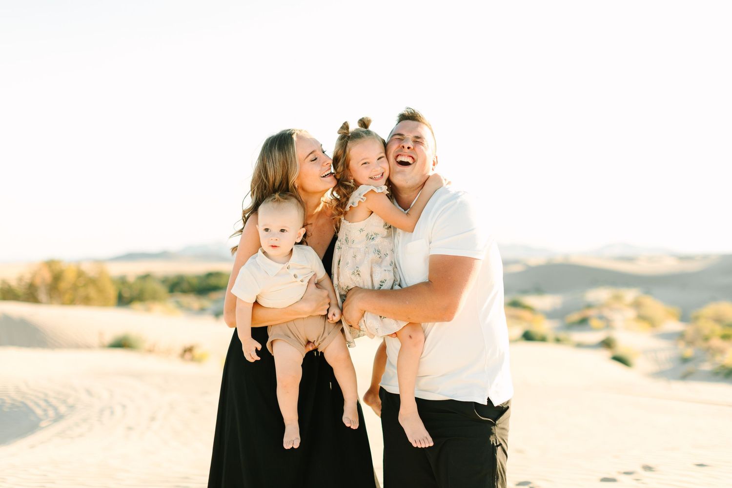 A family wearing neutral colors poses together on a sandy beach at sunset, creating a warm and intimate portrait.