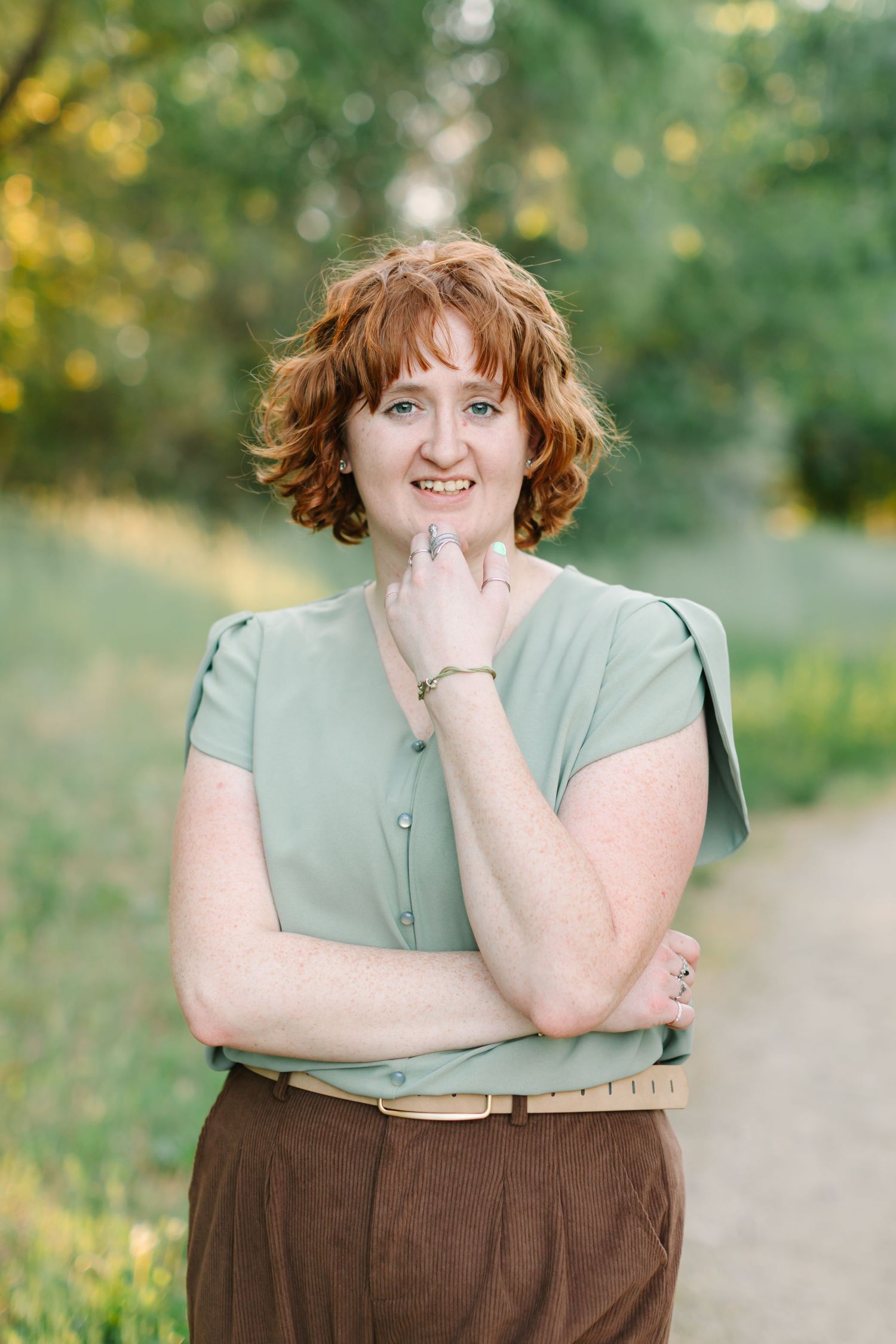 A person in a sage green top and brown skirt stands on a path with a natural blurred background.