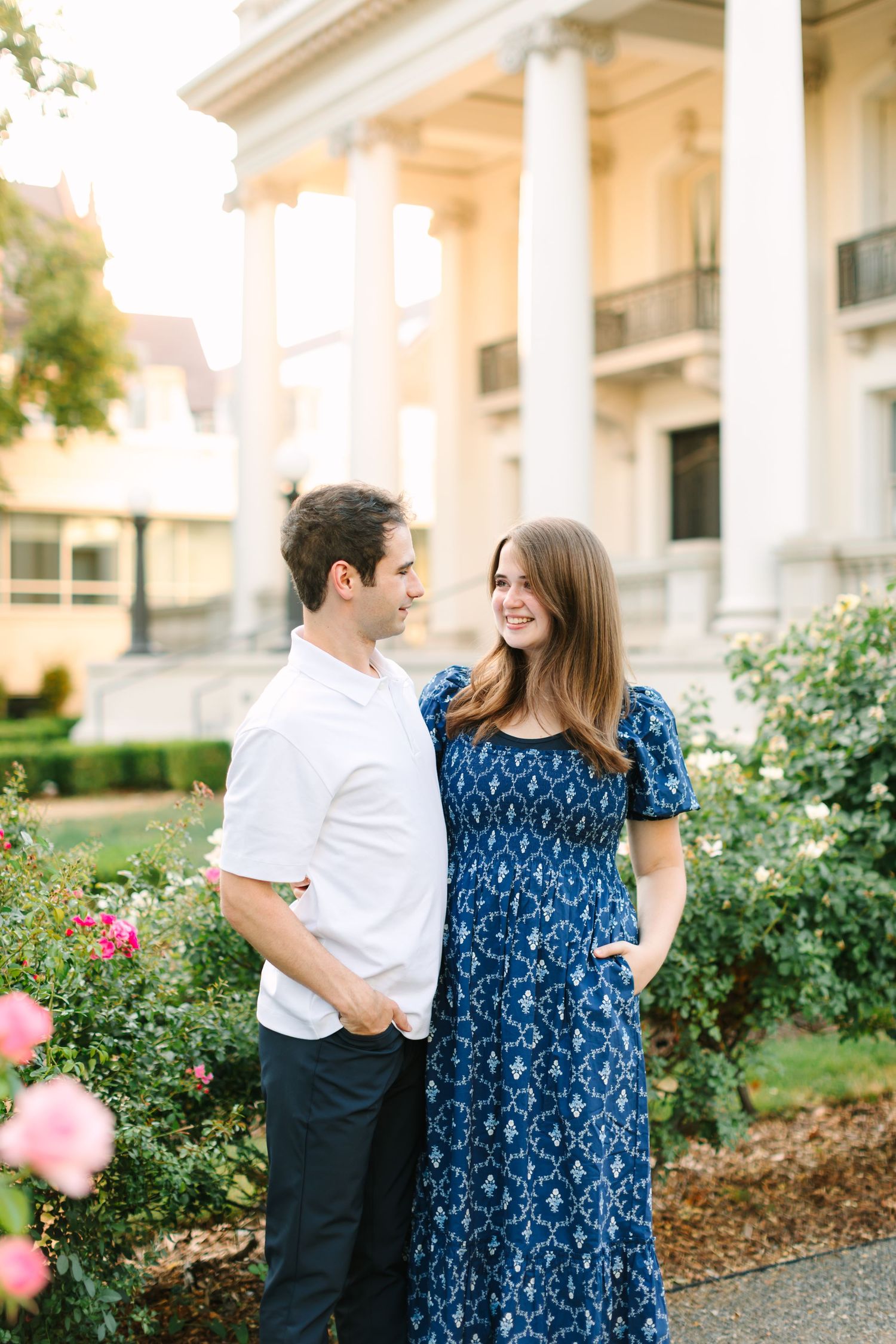 A couple smiles at each other in a garden, woman in blue patterned dress, man in white shirt, elegant white columns behind them.