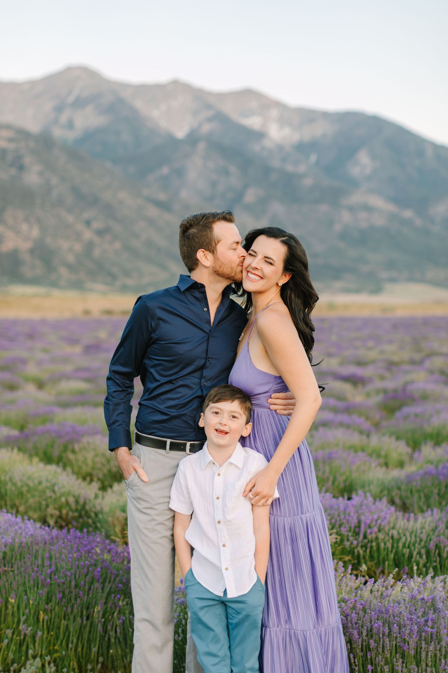 A family poses together in a beautiful lavender field with mountains in the background during sunset.
