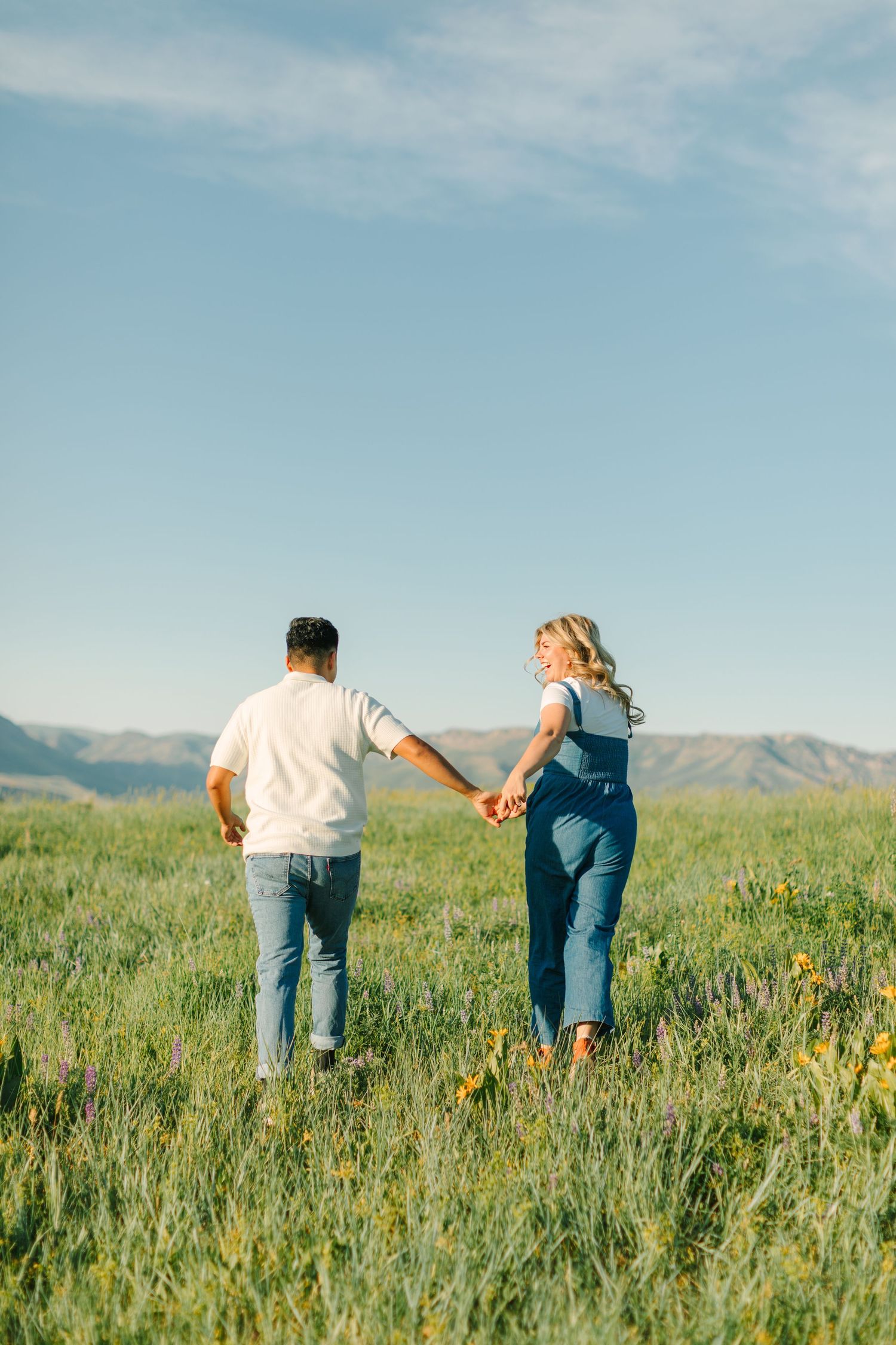 A couple walks hand in hand through a scenic wildflower meadow on a sunny day in Logan, Utah for their engagement photoshoot.