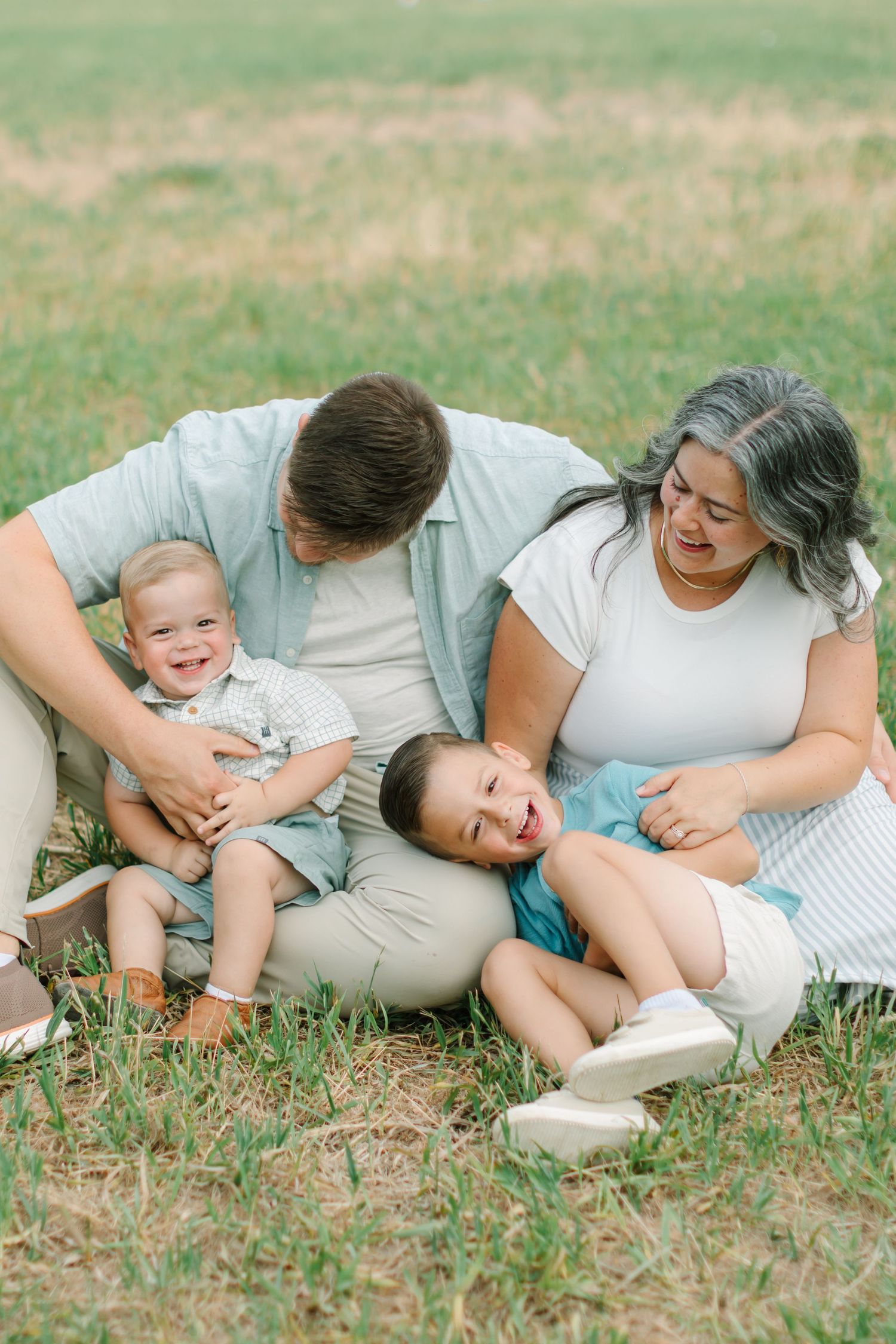 Family laying together in grass enjoying playful candid moments during outdoor photo session.