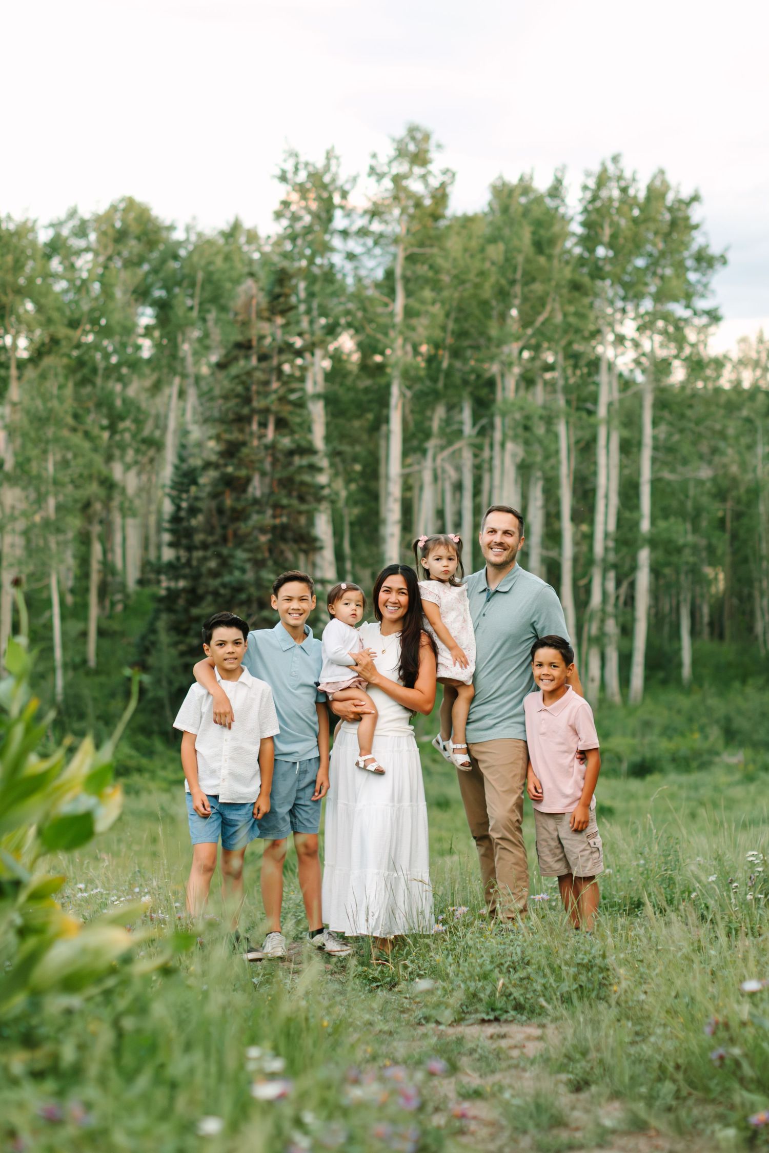 Family of seven posing outdoors in a lush green aspen forest, wearing coordinated pastel outfits for a portrait in Heber, Utah.