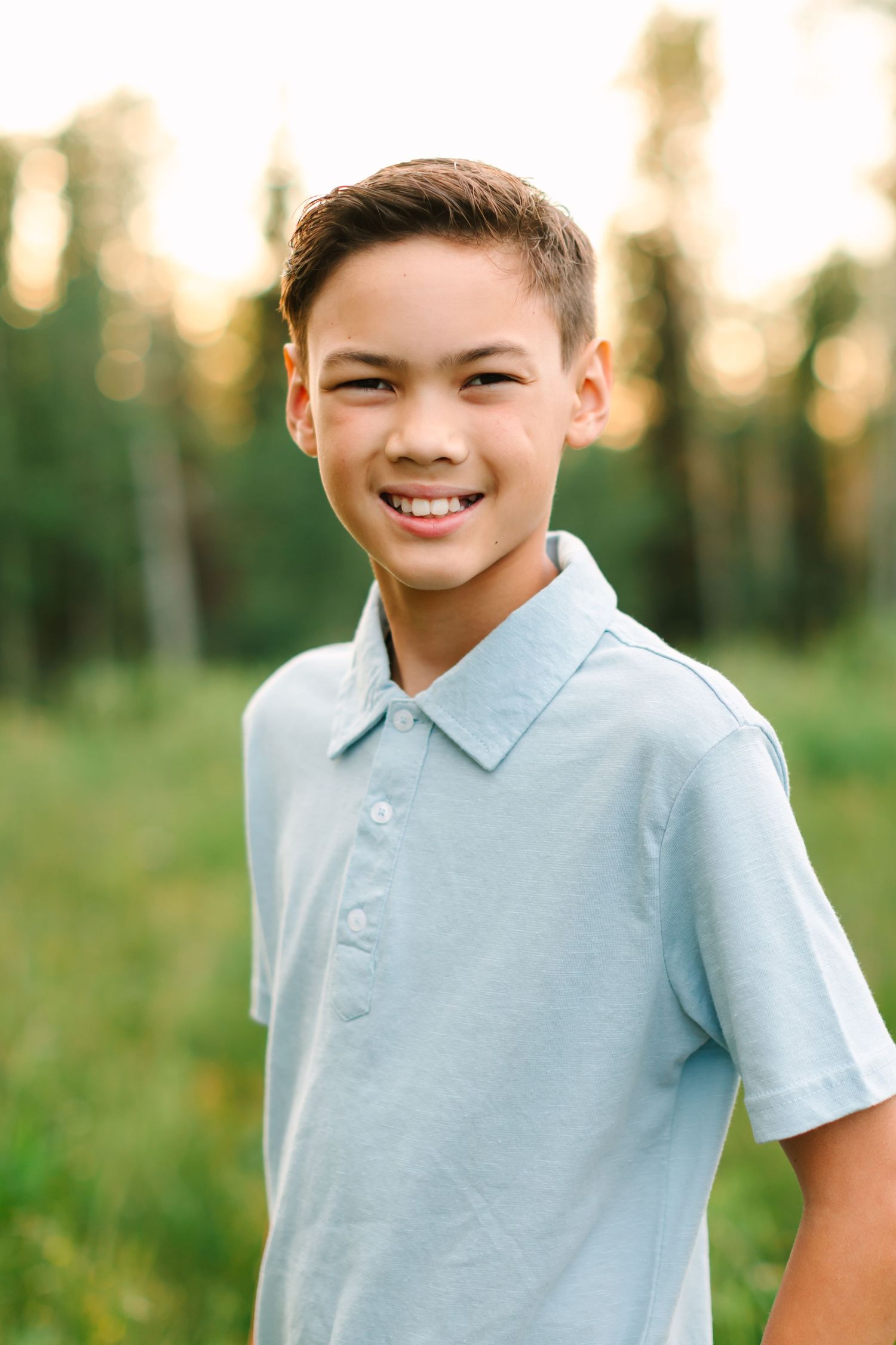 Smiling preteen boy in light blue polo shirt standing outdoors in a sunny green field with trees in background.