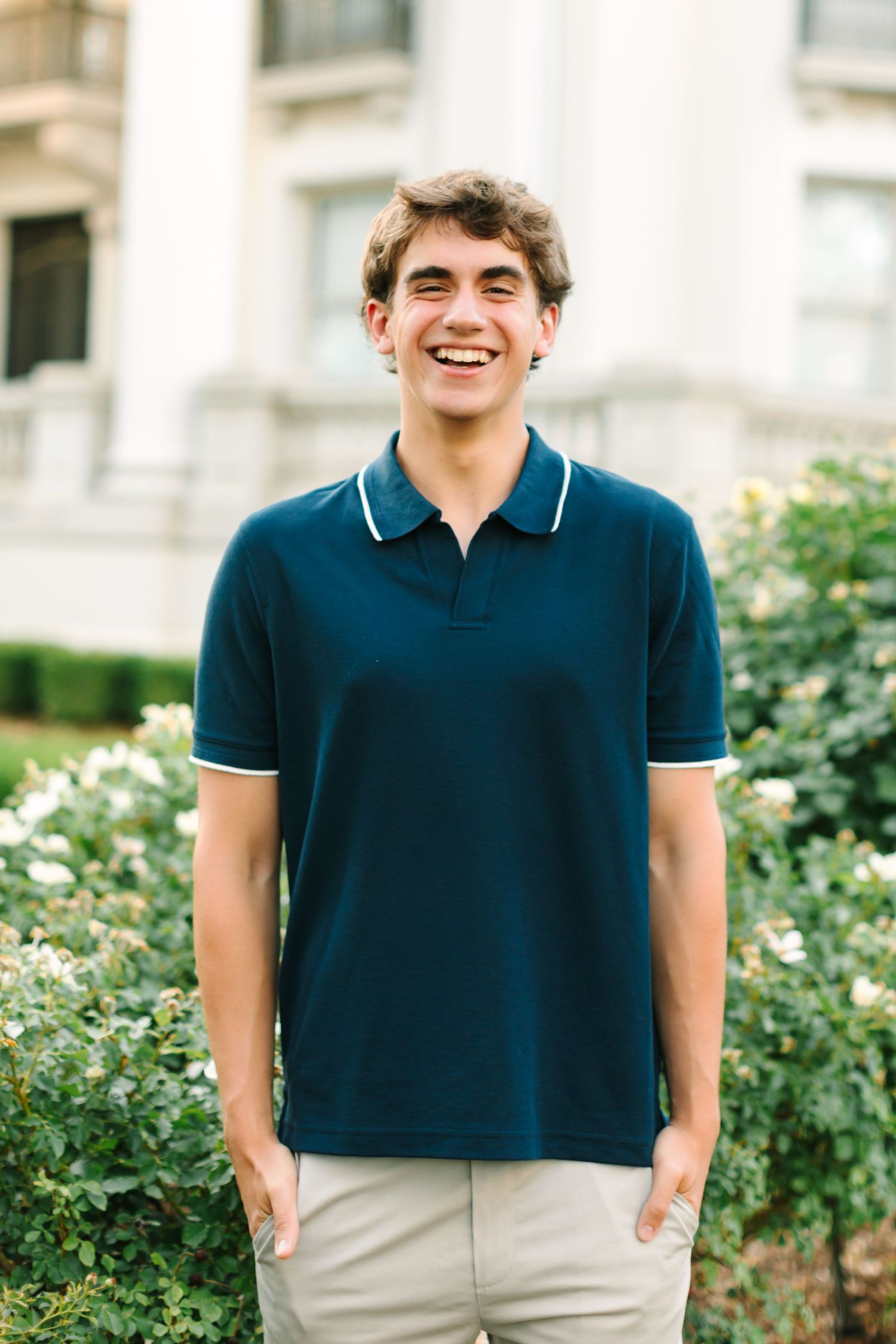 Smiling young man in navy polo shirt standing outdoors near white flowering bushes and a white building.