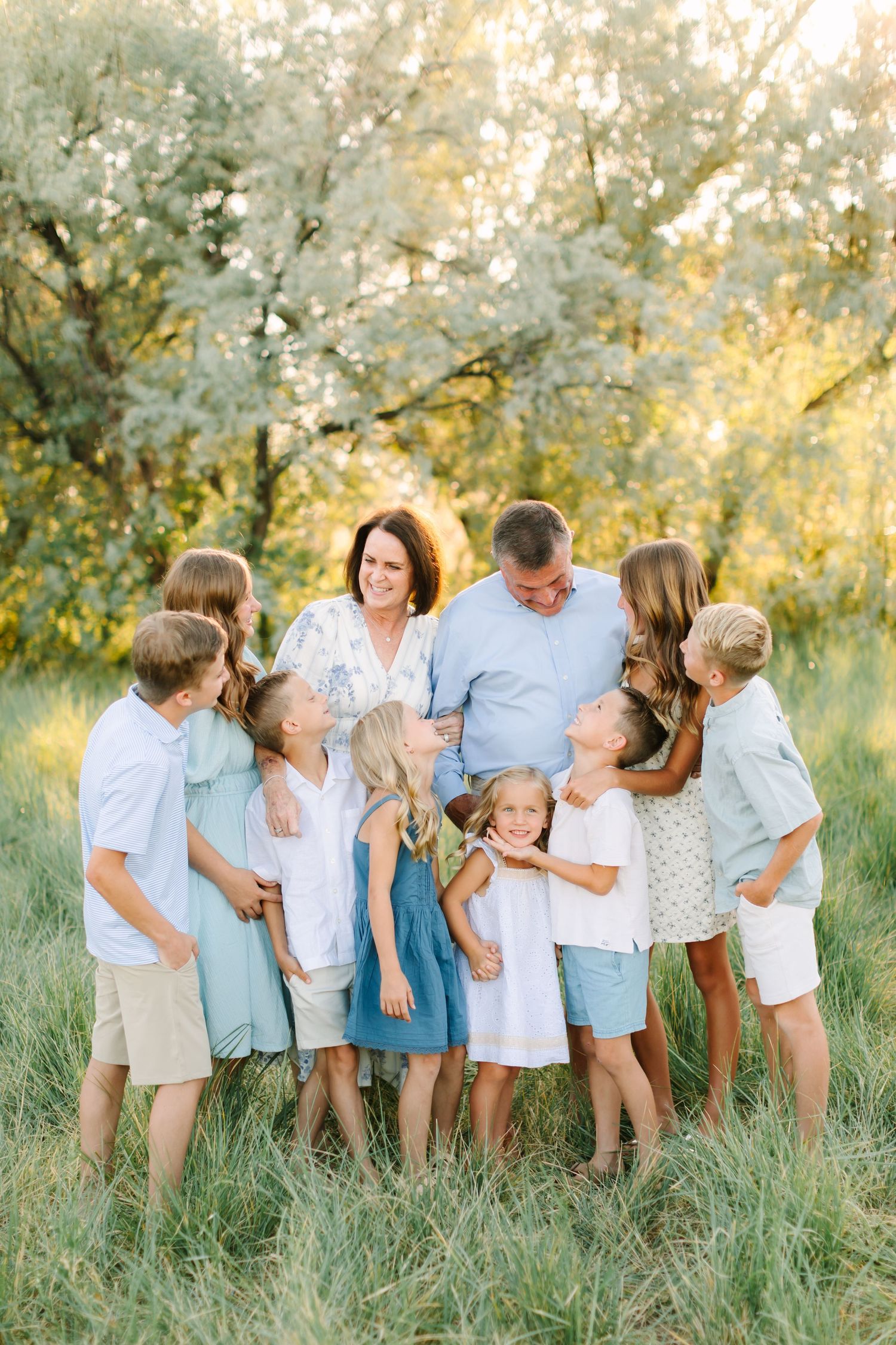 Large family with two parents and seven children posing together in a sunny green field with blooming trees.