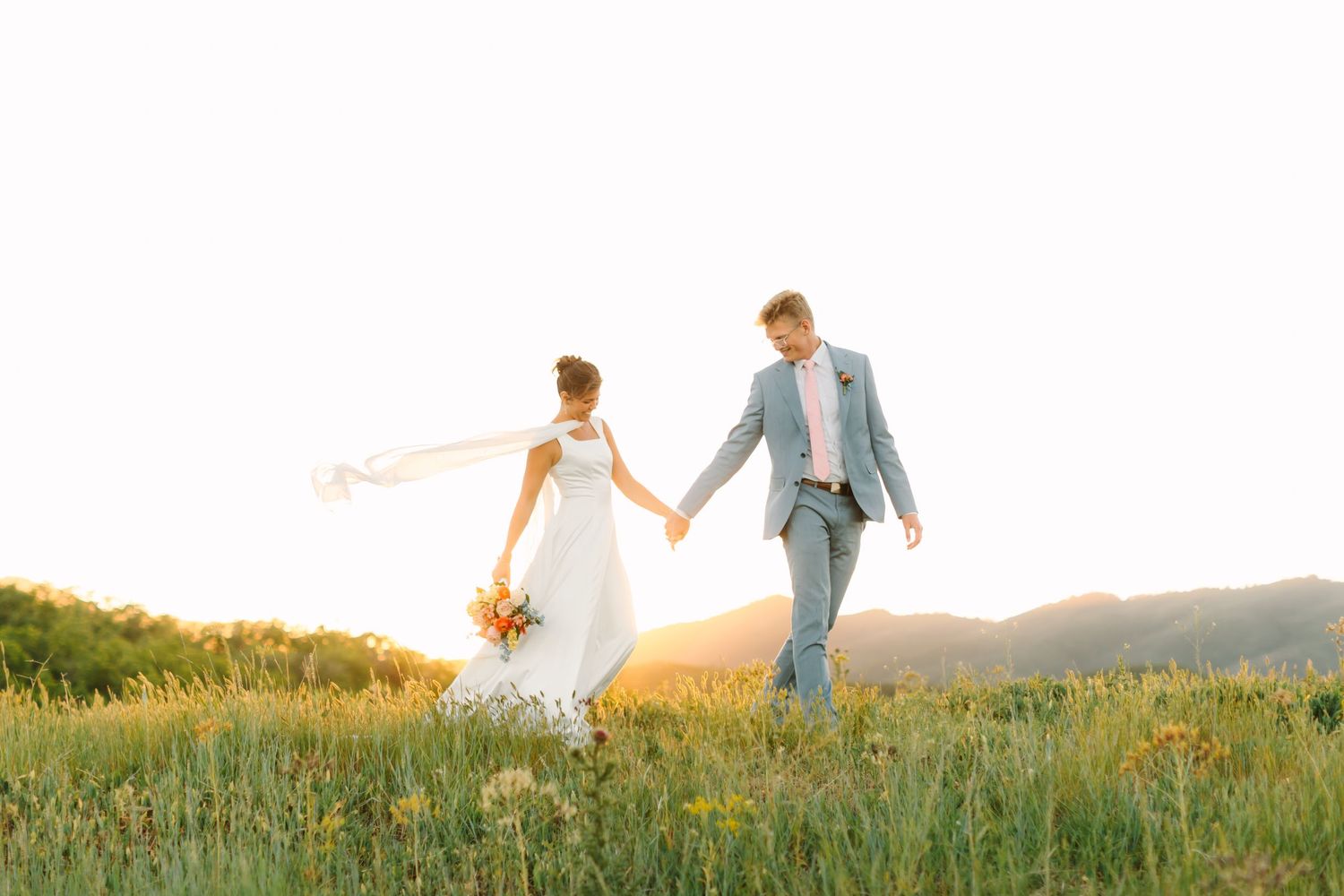 Bride and groom holding hands walking through a sunlit mountain meadow, bride in white gown holding colorful bouquet.