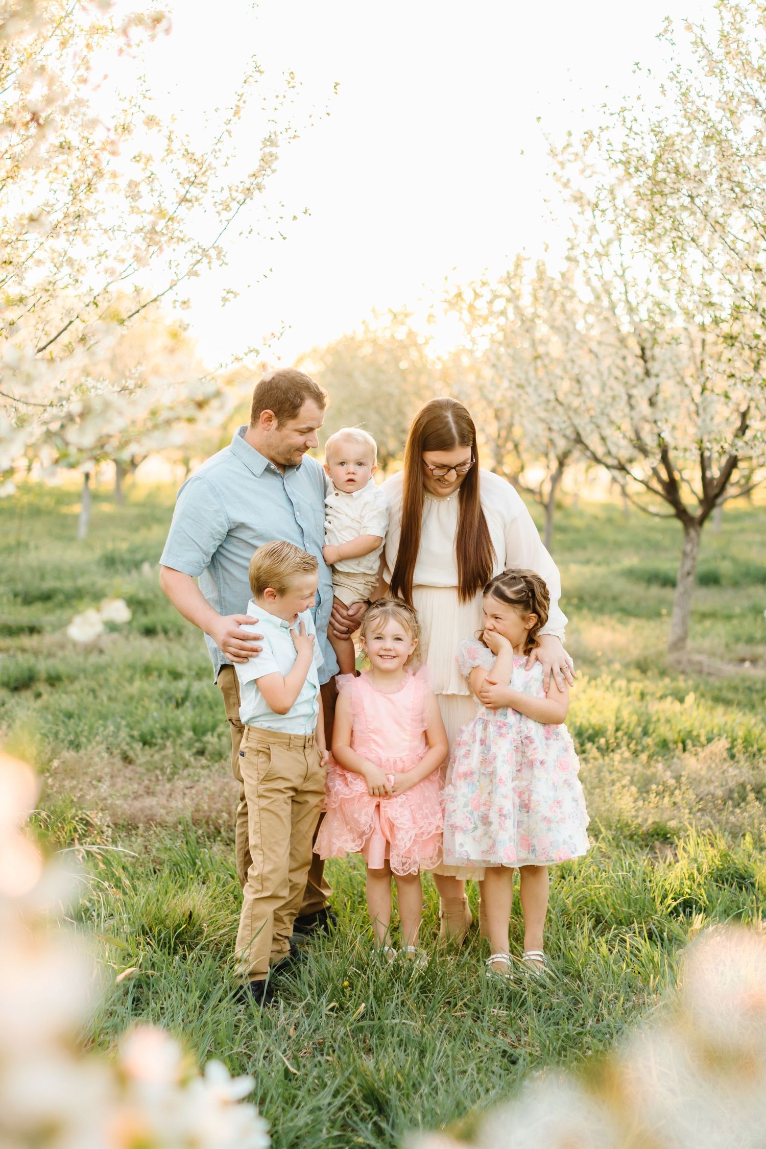 Family of six poses in blooming spring orchard at golden hour, parents with four young children in pastel outfits.
