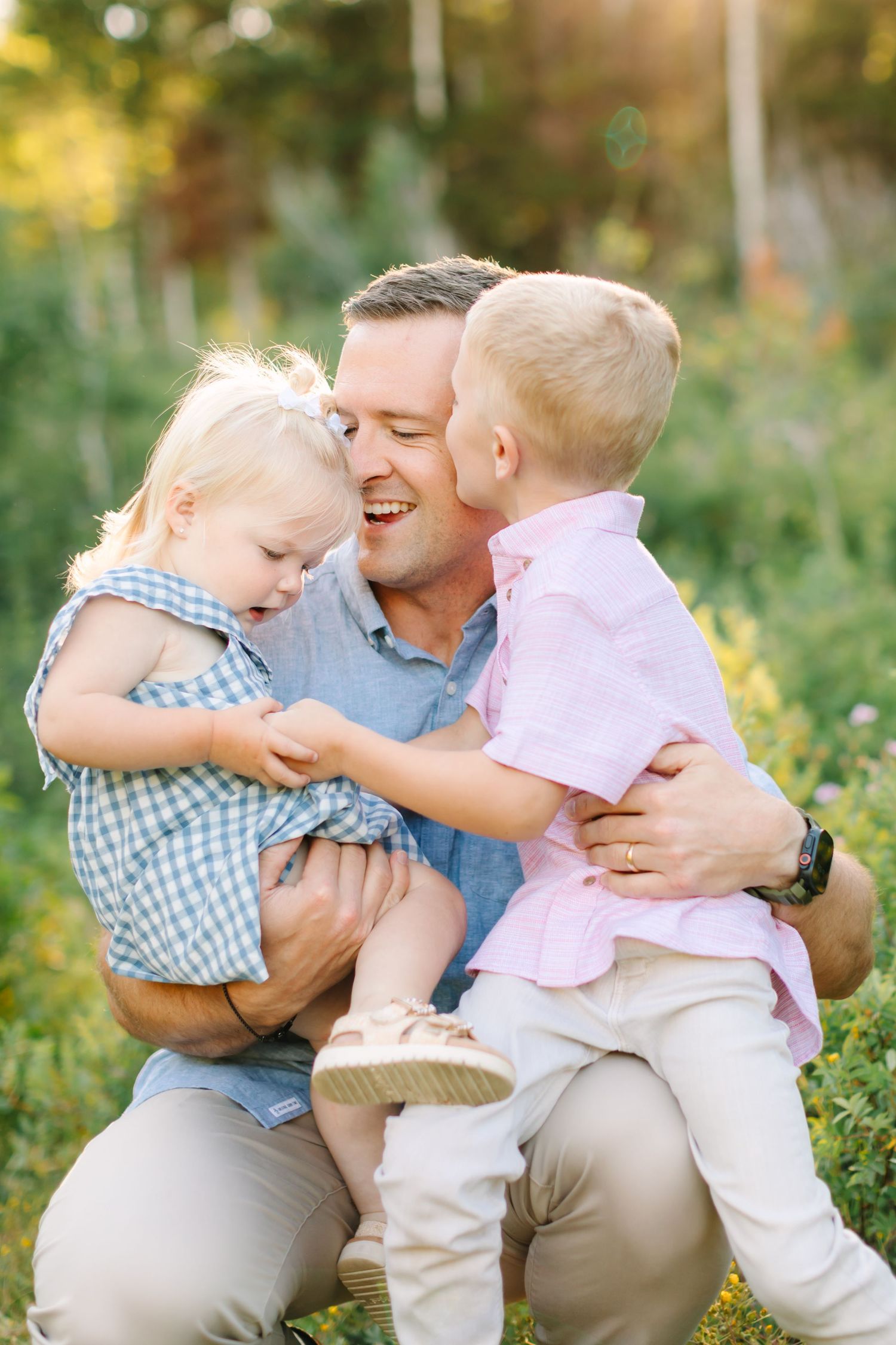 A smiling father crouches outdoors holding two young blonde children, a toddler girl and a boy, in a sunny green field in American Fork, UT.