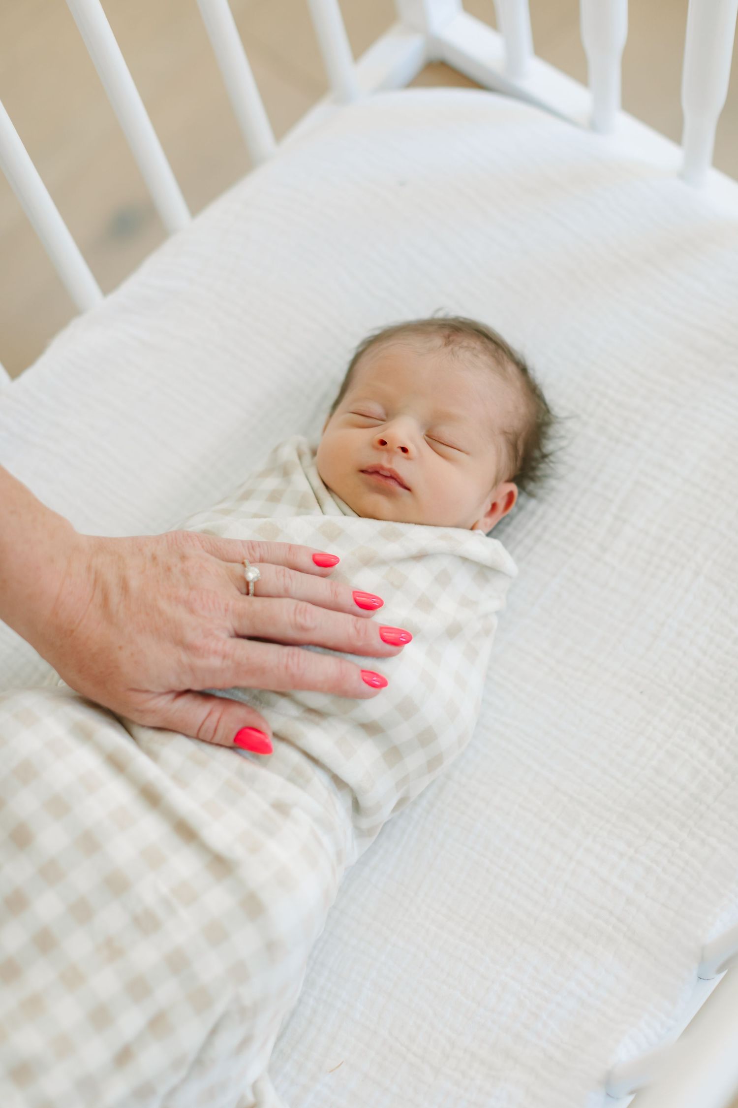 A sleeping newborn baby in white clothing rests peacefully on white bedding with a hand gently touching.