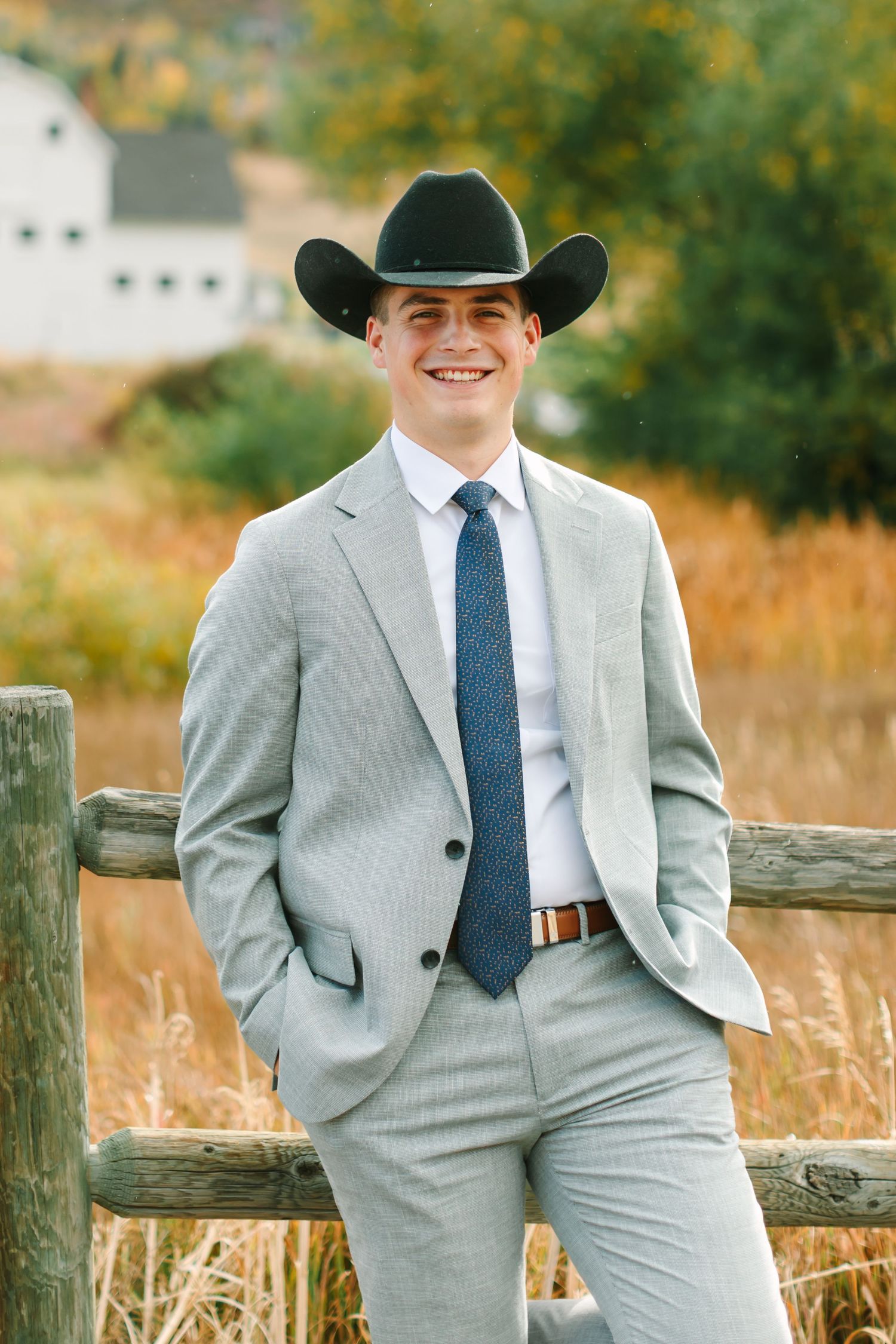Professional western-style portrait session in a rustic setting with light gray suit and cowboy hat against autumn field.