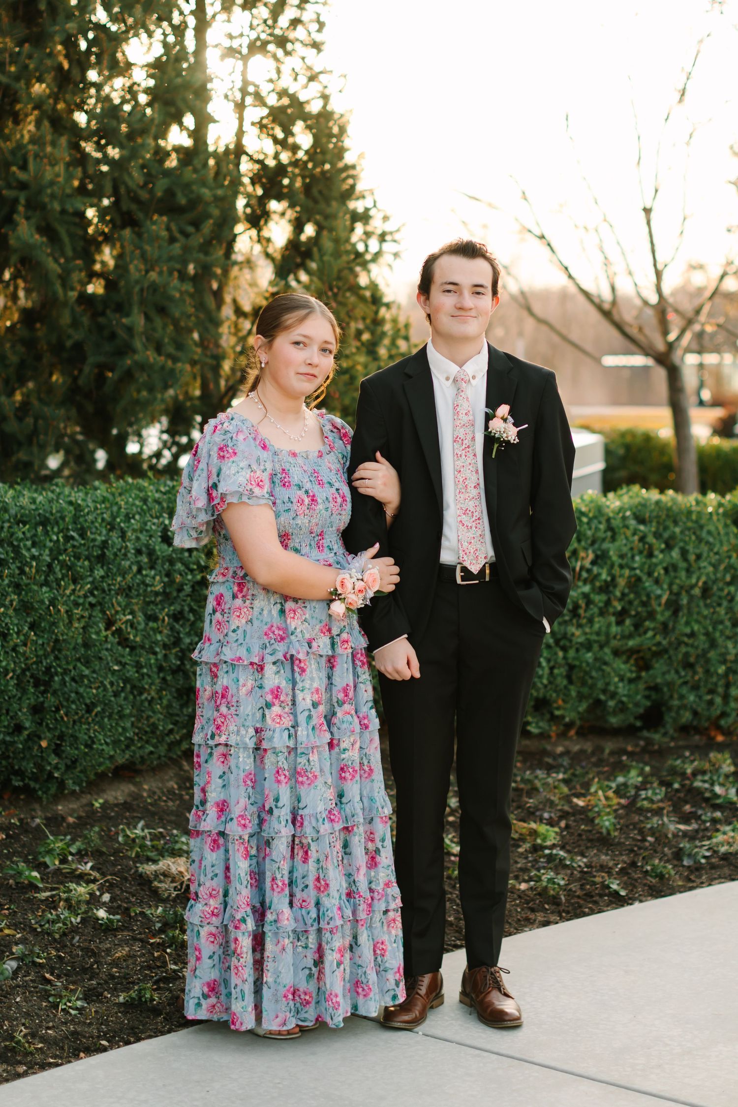 Couple in formal attire pose together against green hedge backdrop during golden hour.