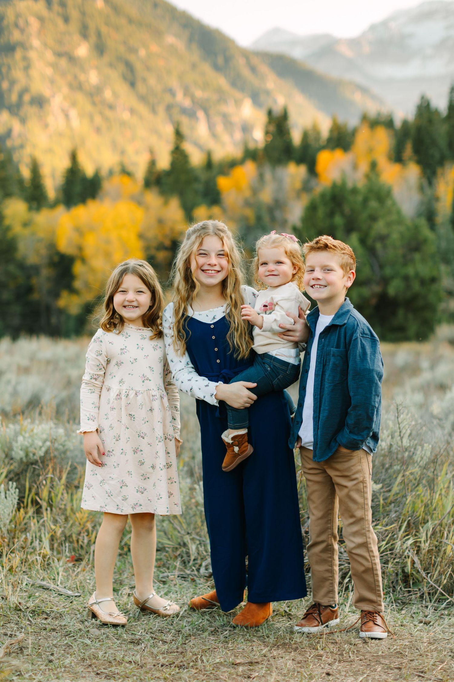 Children in coordinated outfits stand together against a backdrop of autumn mountains and golden foliage.