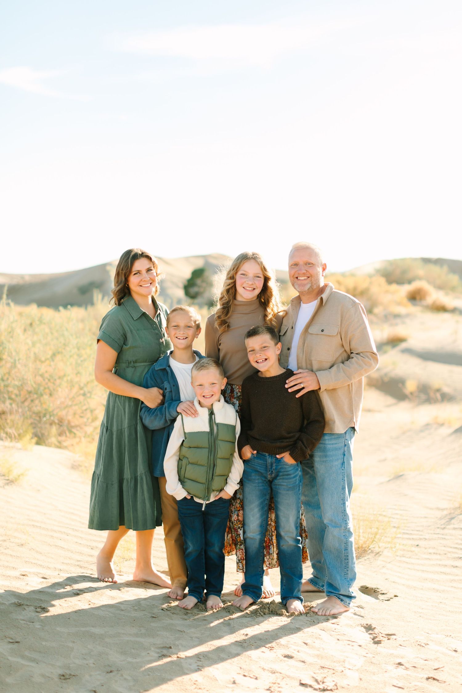 A family poses together on sandy dunes during golden hour in casual outdoor attire.