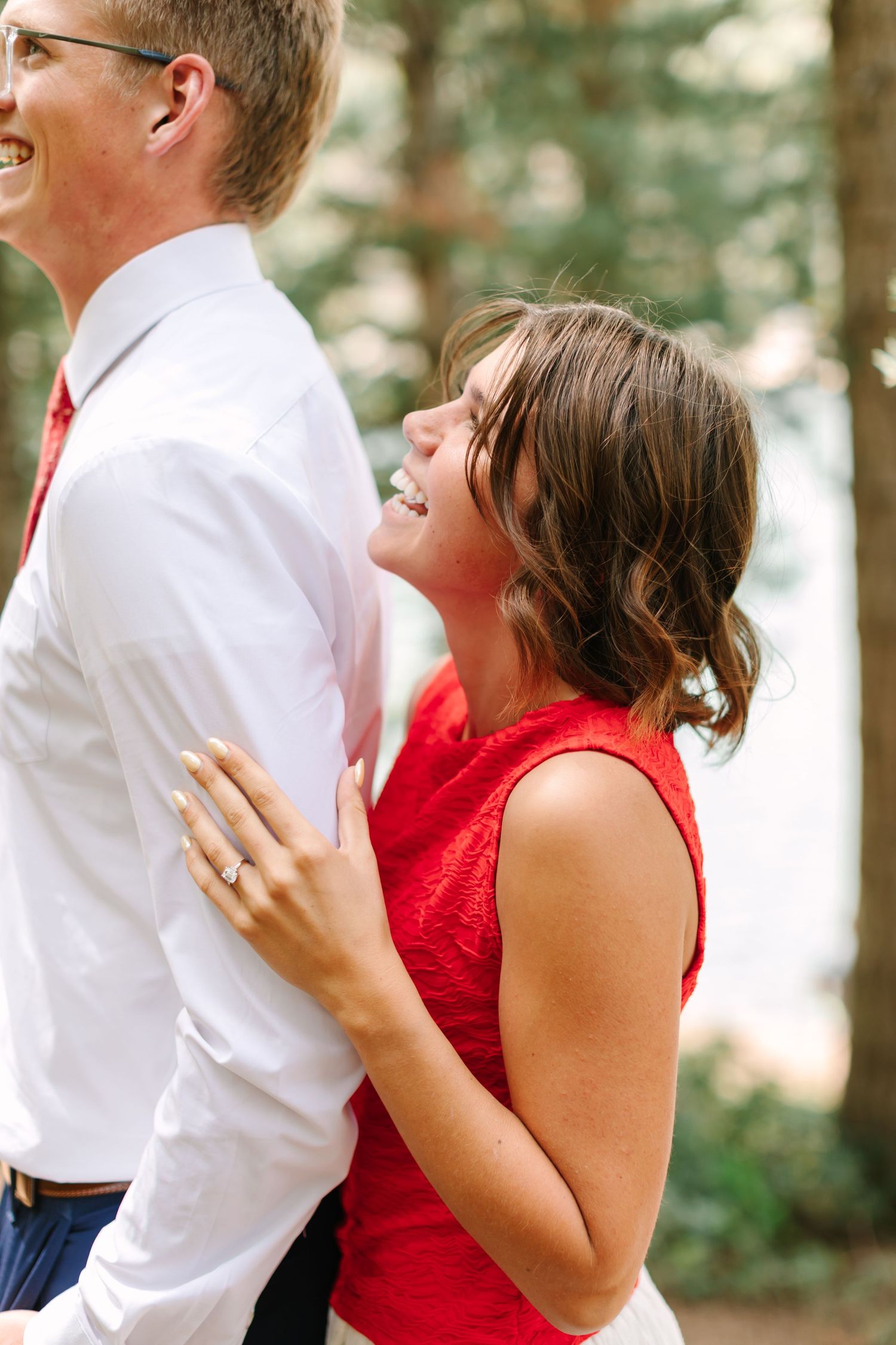 A laughing woman in a red dress embraces a man in a white shirt outdoors among trees, wearing an engagement ring.